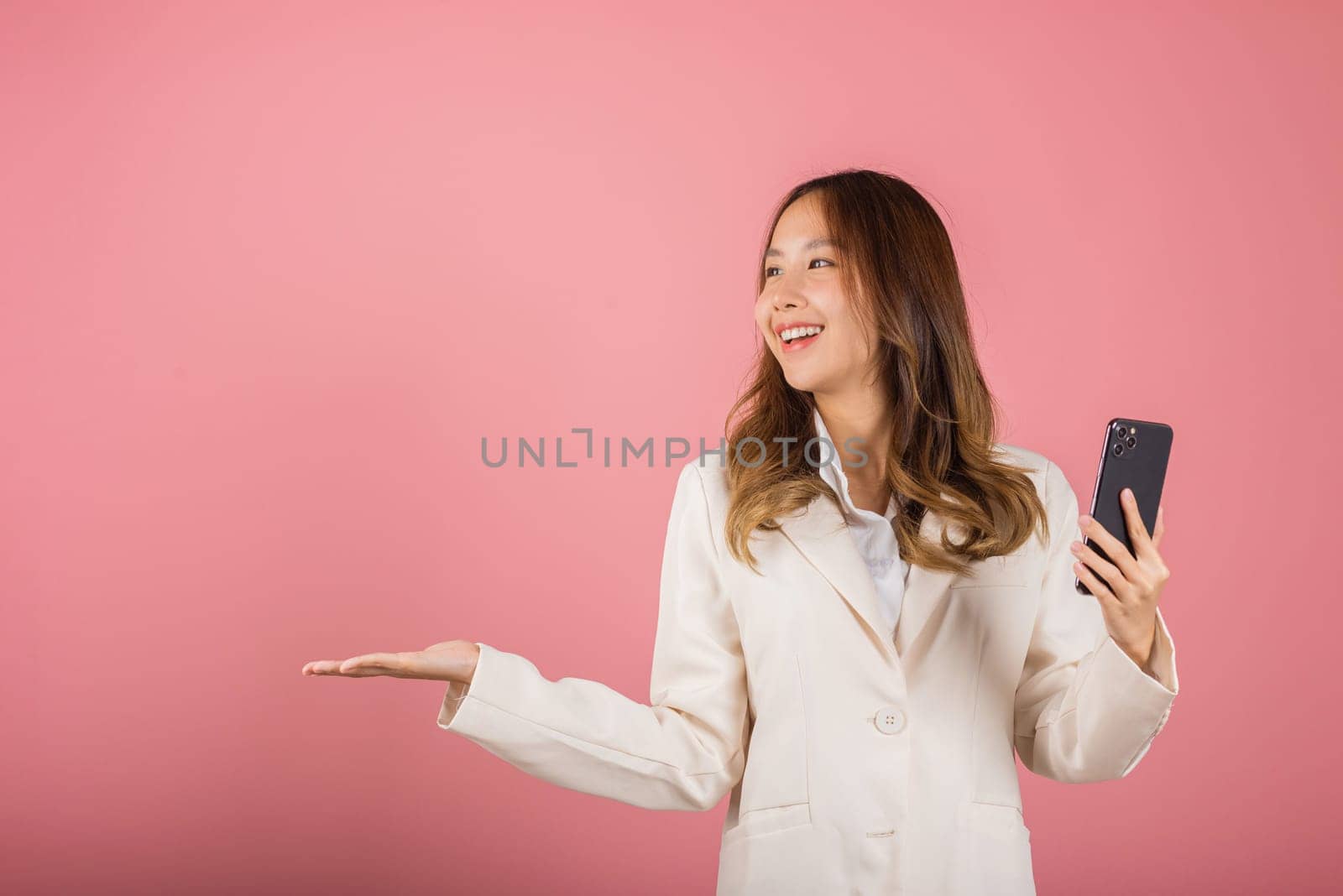 Happy Asian portrait beautiful cute young woman excited holding mobile phone blank screen presenting product with palm of hand at empty, studio shot isolated on pink background, female look to space