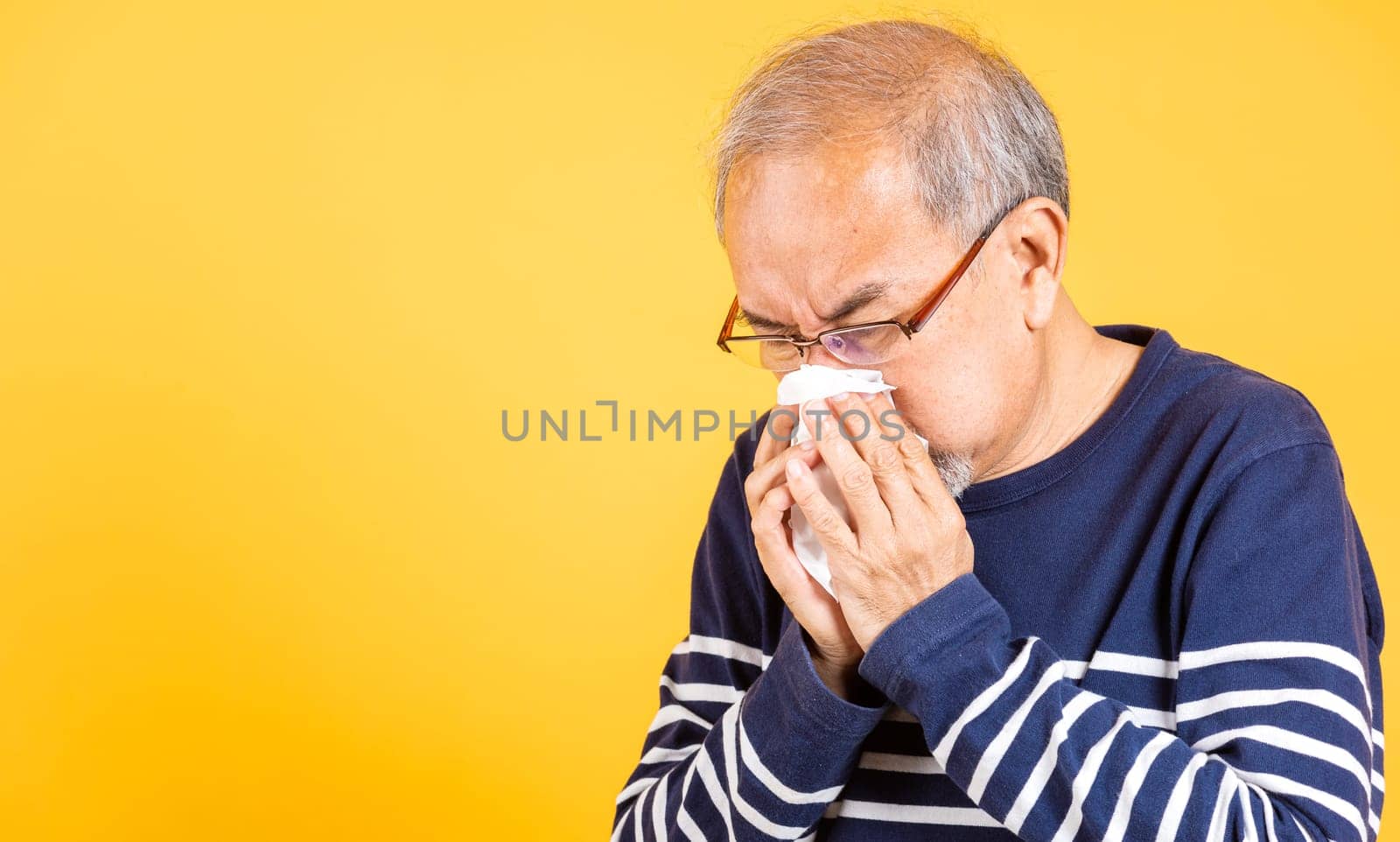 Portrait senior old man blowing nose with tissue studio shot isolated on yellow background, Asian elder man cold having flu and sneezing from sickness virus problem use tissue, pensioner unwell