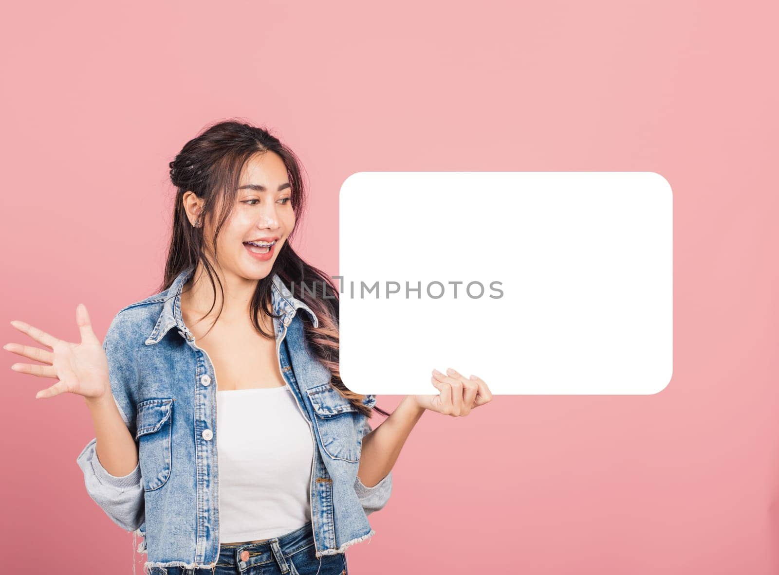 Happy Asian beautiful young woman smiling excited wear denims holding empty speech bubble sign, Portrait female posing show up for your idea looking at bubble, studio shot isolated on pink background