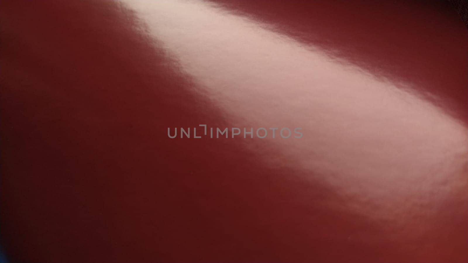 A long flare is a visage of light on shiny burgundy paper. Light spot on a dark red surface that reflects light. Abstract monochrome background. Strip from top left to right down.
