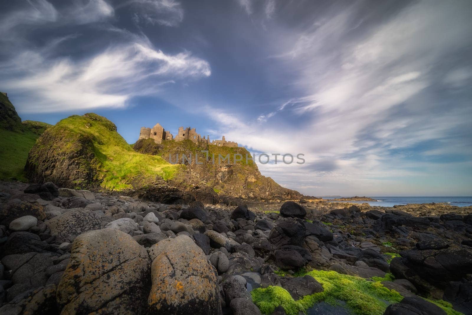 Dunluce Castle seen from behind of mossy rocks on a shore, nested on the edge of cliff, part of Wild Atlantic Way, Bushmills, Northern Ireland