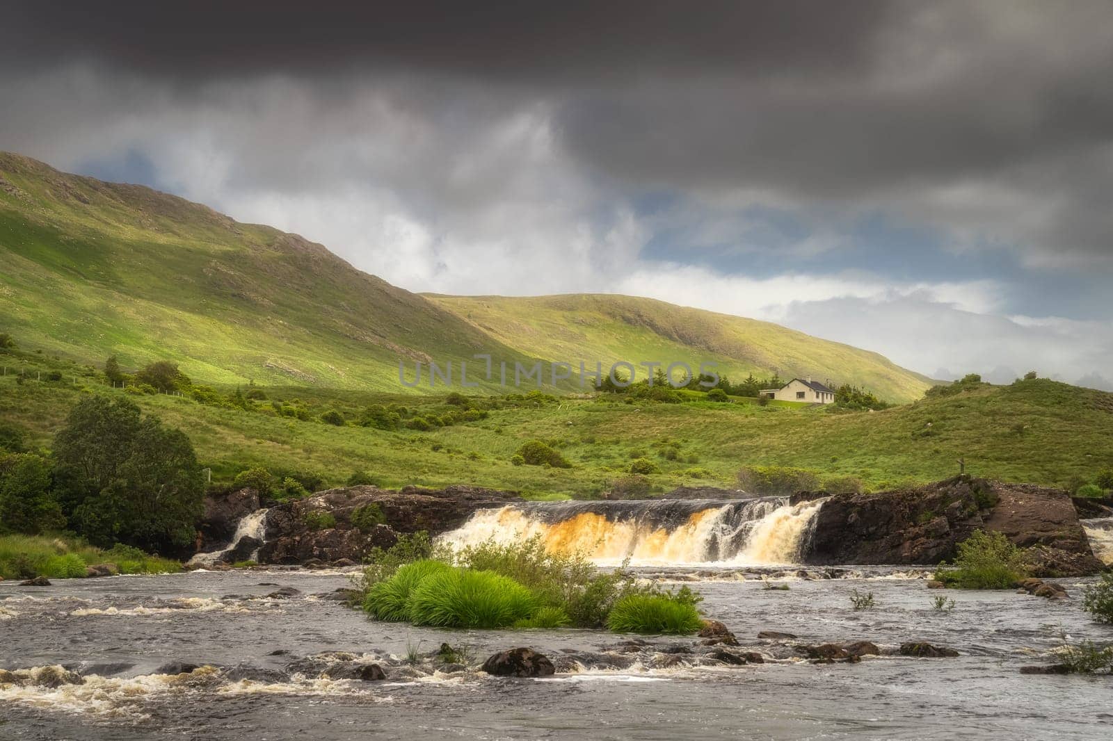 Aasleagh Falls on Erriff River with a single house under Letterass Mountains with adrk dramatic sky in background, County Mayo, Ireland