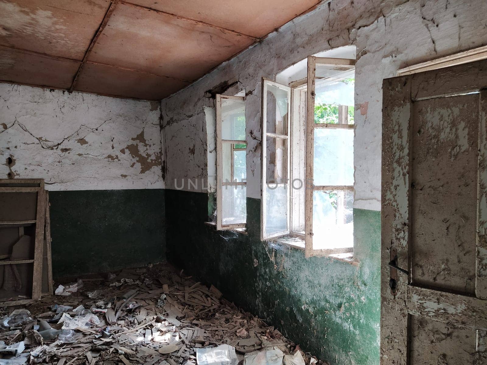 Inside an abandoned house. Scattered trash on the floor. Green paint, cracks and cobwebs on the wall. The white upper half of the wall. Open windows through which sunlight shines through. Old door.
