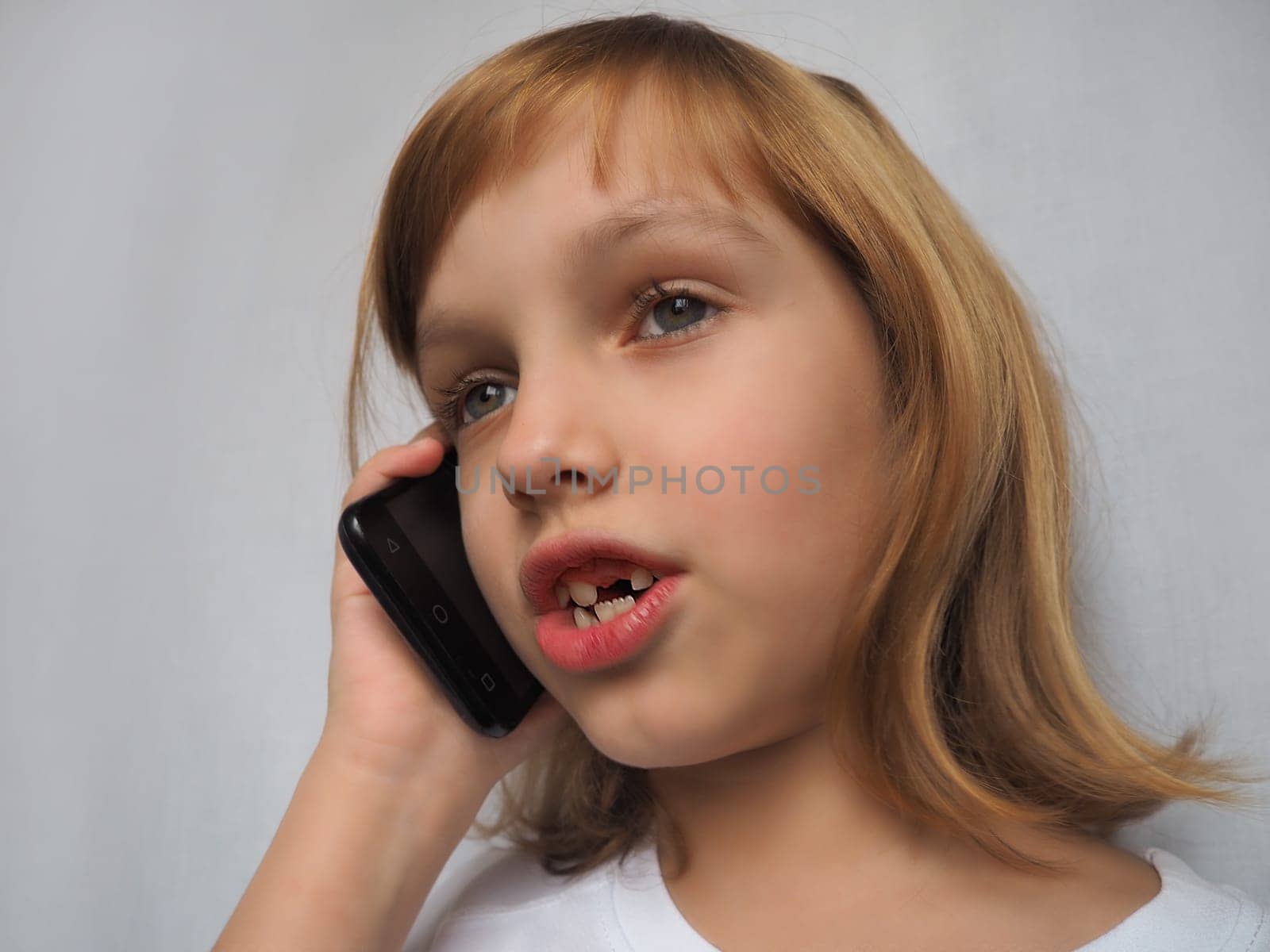 A 7-year-old girl without a front tooth. The child smiles, looks to the side and and talks on the phone or smartphone. Change of milk teeth in childhood. Orthodontics and dental practice.