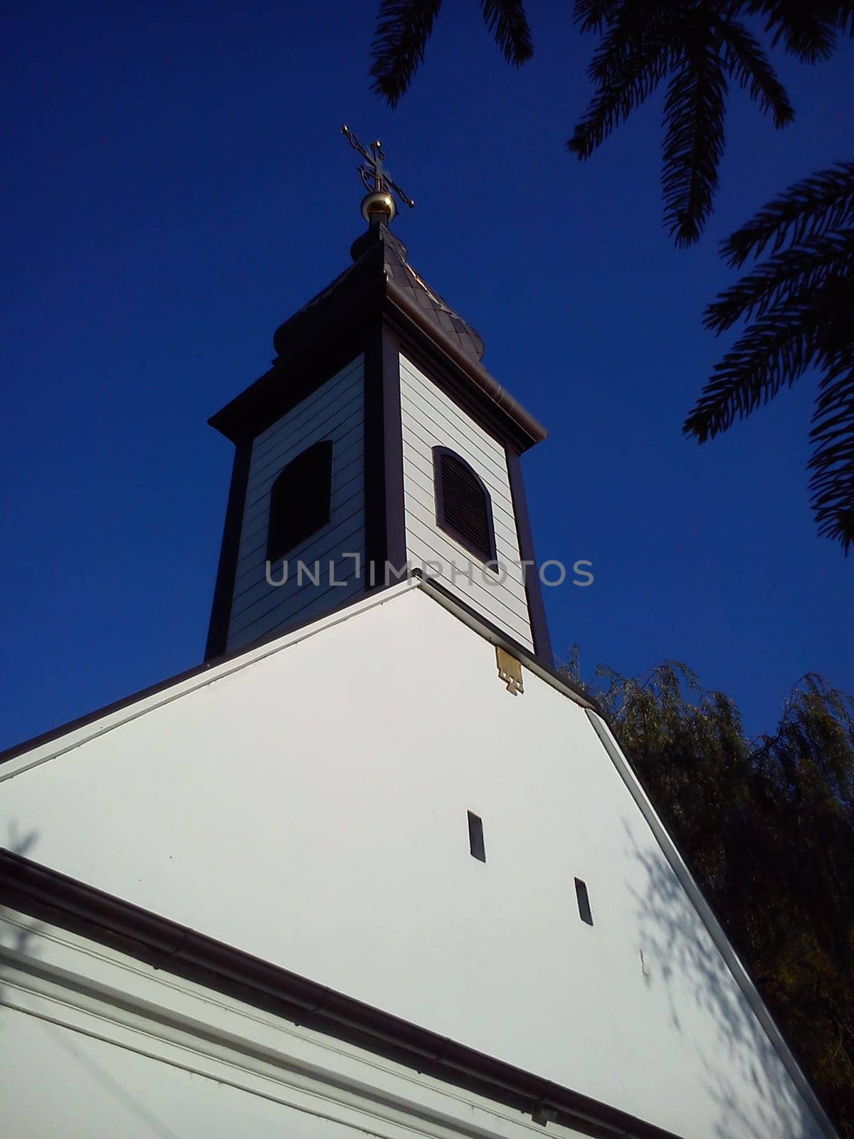 Roof and church dome with a cross. Christian temple, plastered and painted white. The sound of a small wind window and a belfry with a bell. Gilded cross against a blue sky.