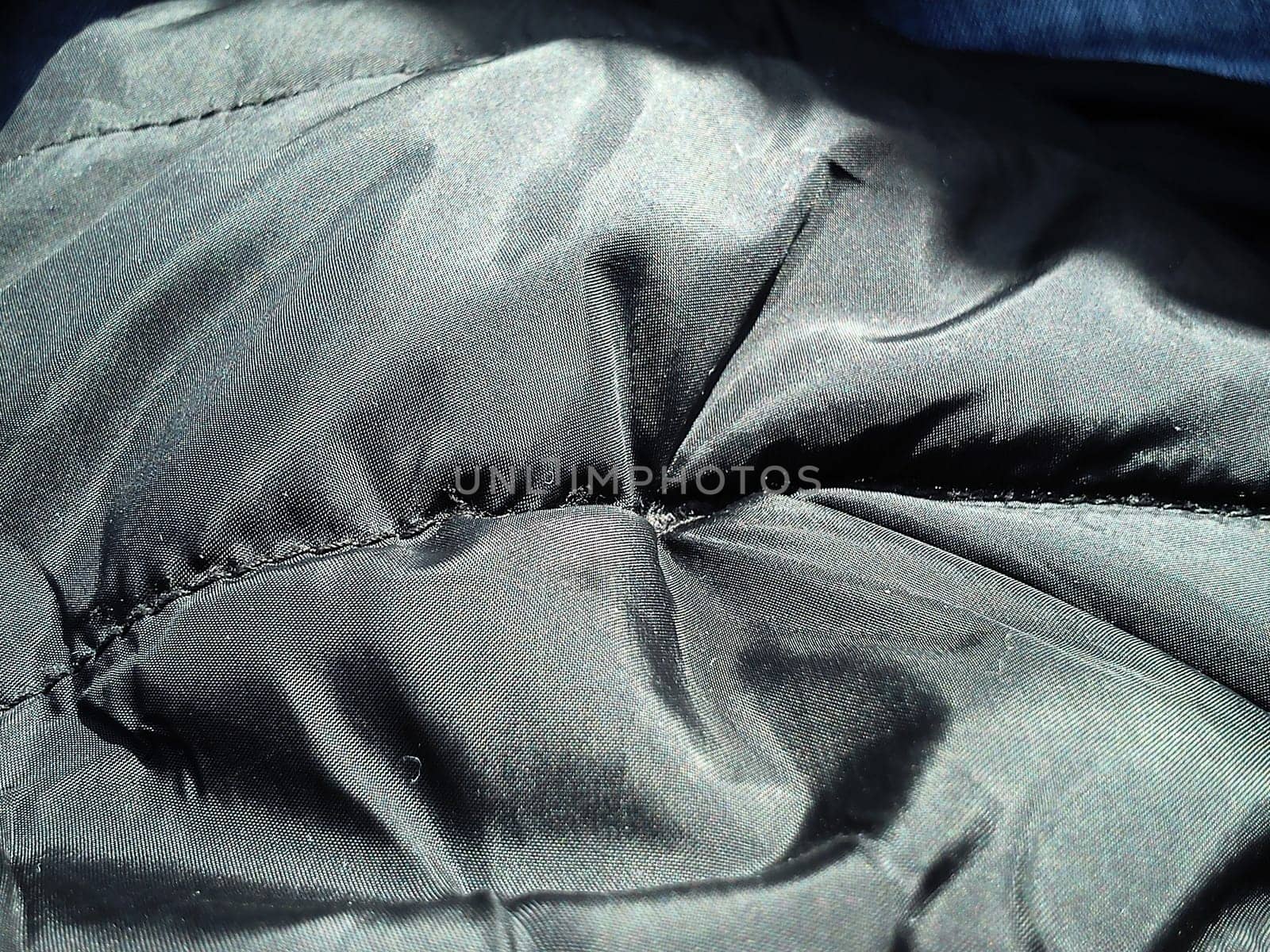 Closeup of black fabric on a winter jacket. Demi-season clothing. Smooth seam with black thread. Slightly shiny synthetic fabric with a seam. Rainproof impregnation. Soft folds. Light and shadow.