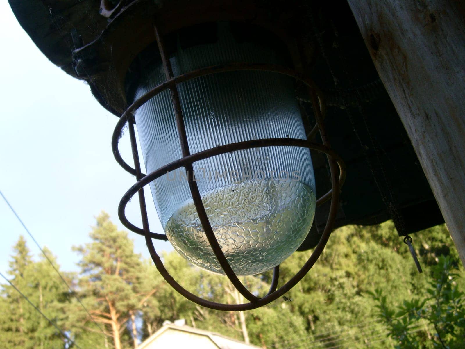 Close up of a street glass lantern in a metal protective vandal proof cage. In the background are visible country houses and green leafy trees