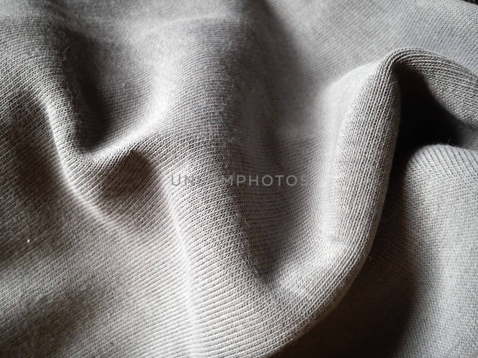 Close-up and texture of a gray knitted fabric, slightly folded in waves or wrinkled. The play of light and shadow on a machine-knitted fabric in natural daylight. Soft, thick, warm cotton material