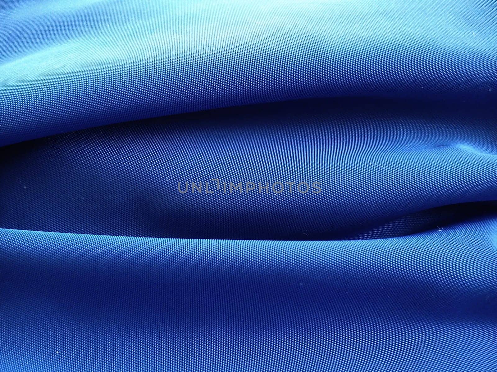 Synthetic fabric in blue, photographed at close range. A noticeable interweaving of threads. Fabric with anti-rain and anti-wind impregnation. A piece of material is laid in waves, light and shadow.