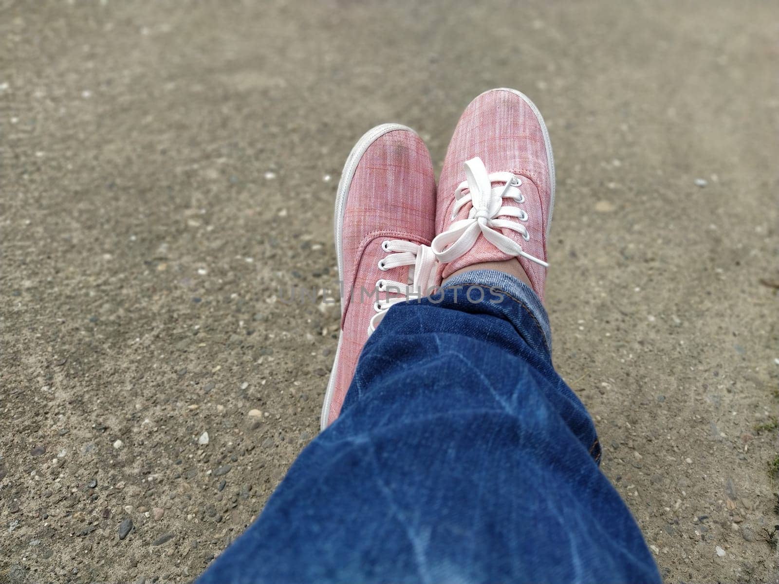 Pink sneakers with white laces on the legs. Blue jeans wrapped underneath. View from above. Carefree and sporty. Crossed legs with a cross. Blurred bokeh. Sand in the background.
