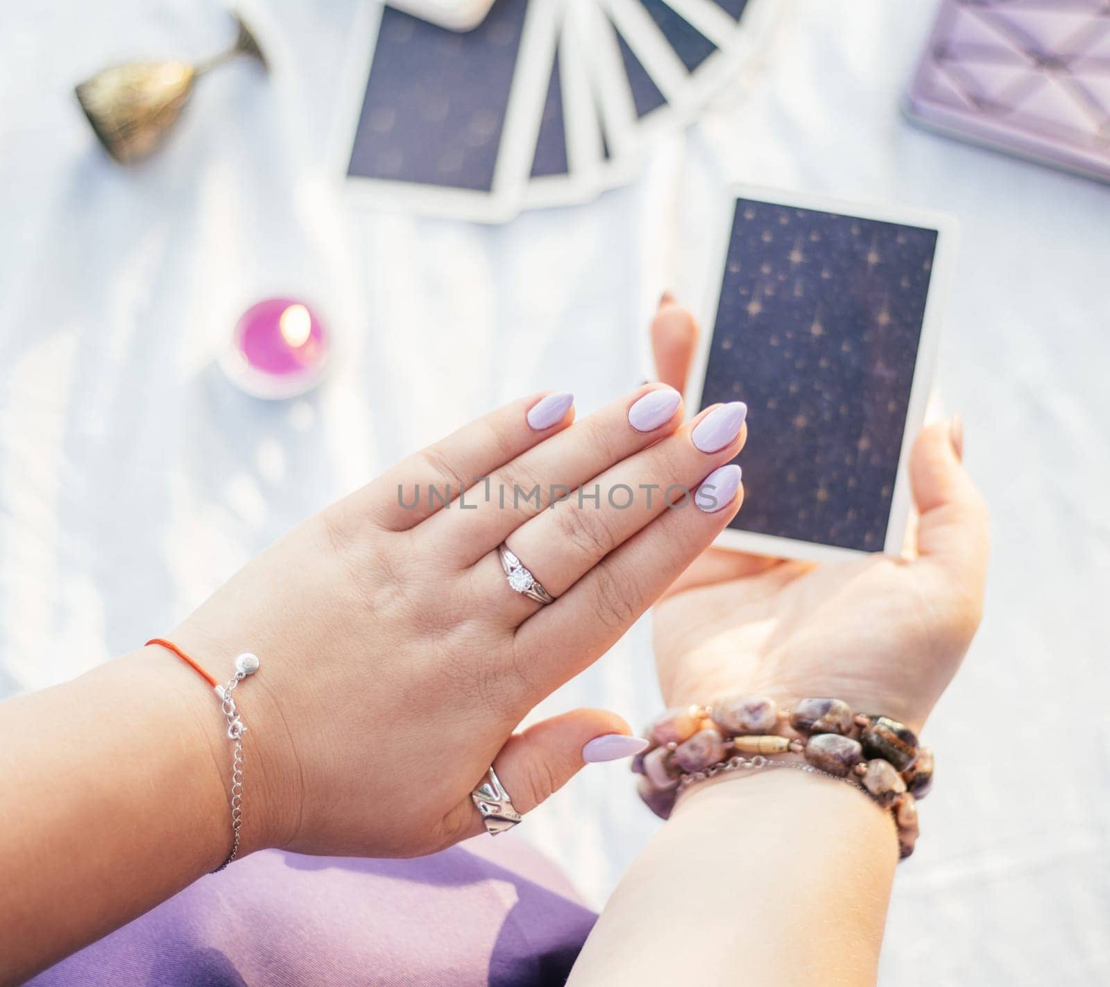 Woman holds her hand with purple nails over Tarot card on white surface with notebook and candle. Top view.