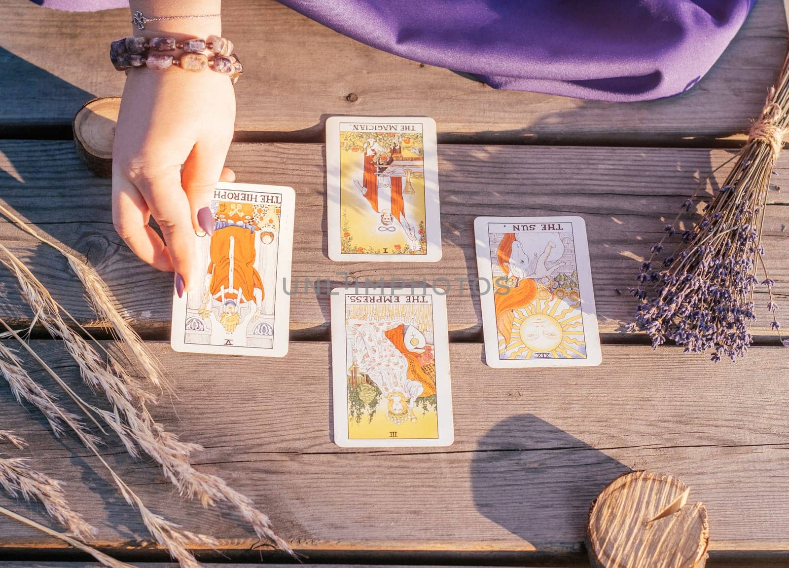 Woman's hand with purple nails lays out one of four Tarot cards spread out on wooden surface next to spikelets and lavender. Minsk, Belarus - 07.28.2021