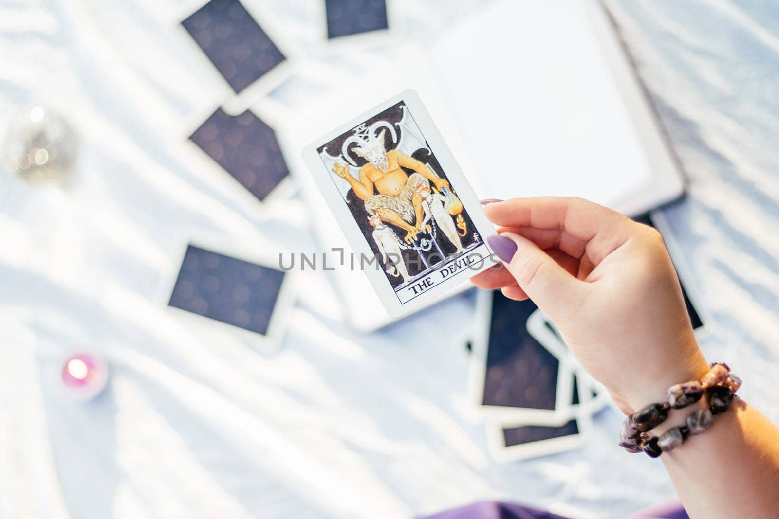 Female hand with purple nails holds tarot card named The Devil over white surface with open notebook and candle. Top view. Minsk, Belarus - 07.28.2021