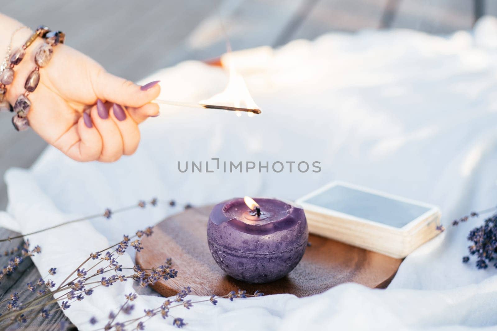 Woman's hand in bracelet with purple long nails lights candle with match, next to deck of cards and lavender flowers on white and wooden surface