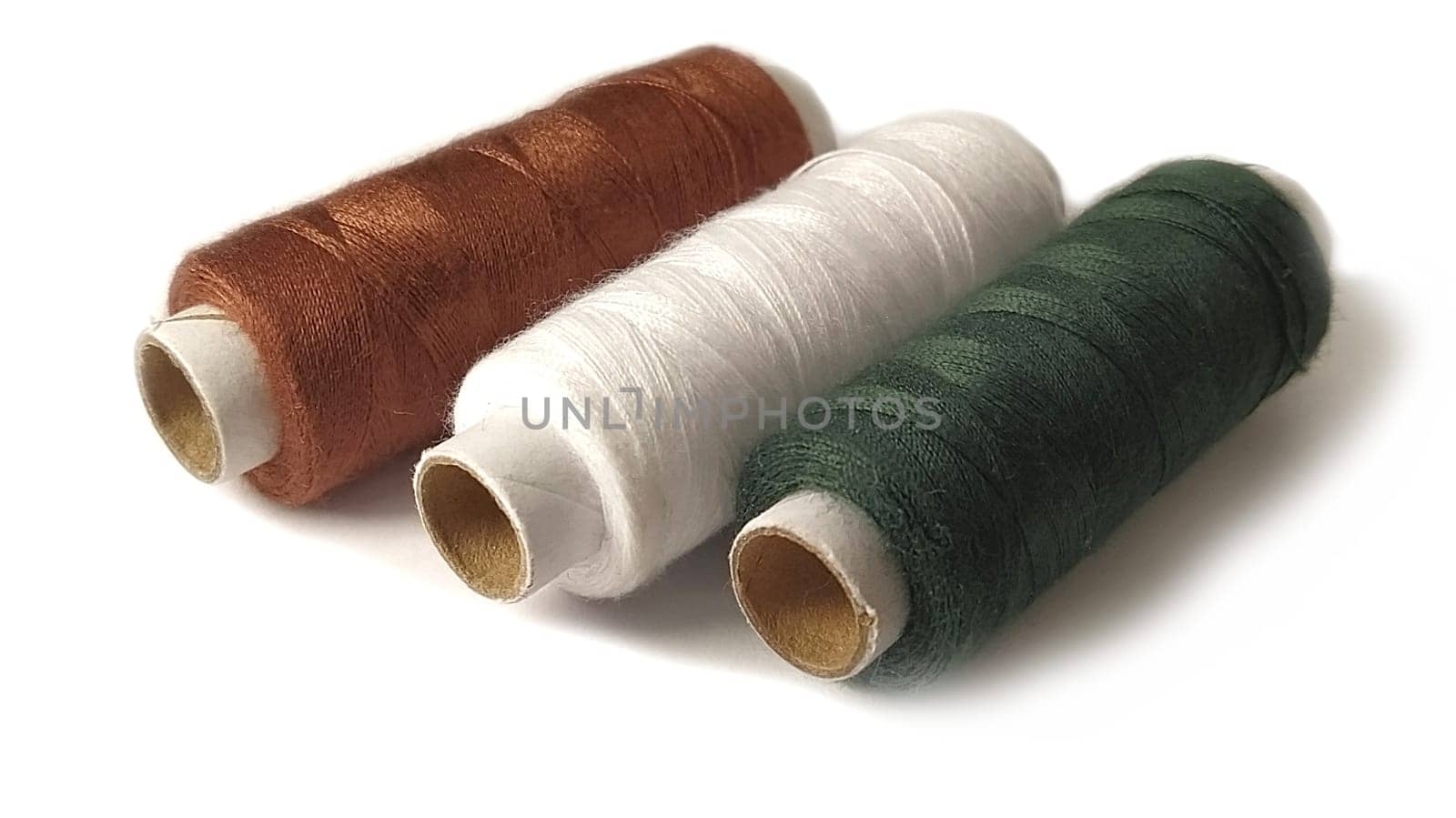 Bobbins and spools of thread of different colors - brown, white and green. Close-up. Natural light on the left side. Isolate on a white background with a light shadow. Skeins of thread. Sewing supplies. Textile production
