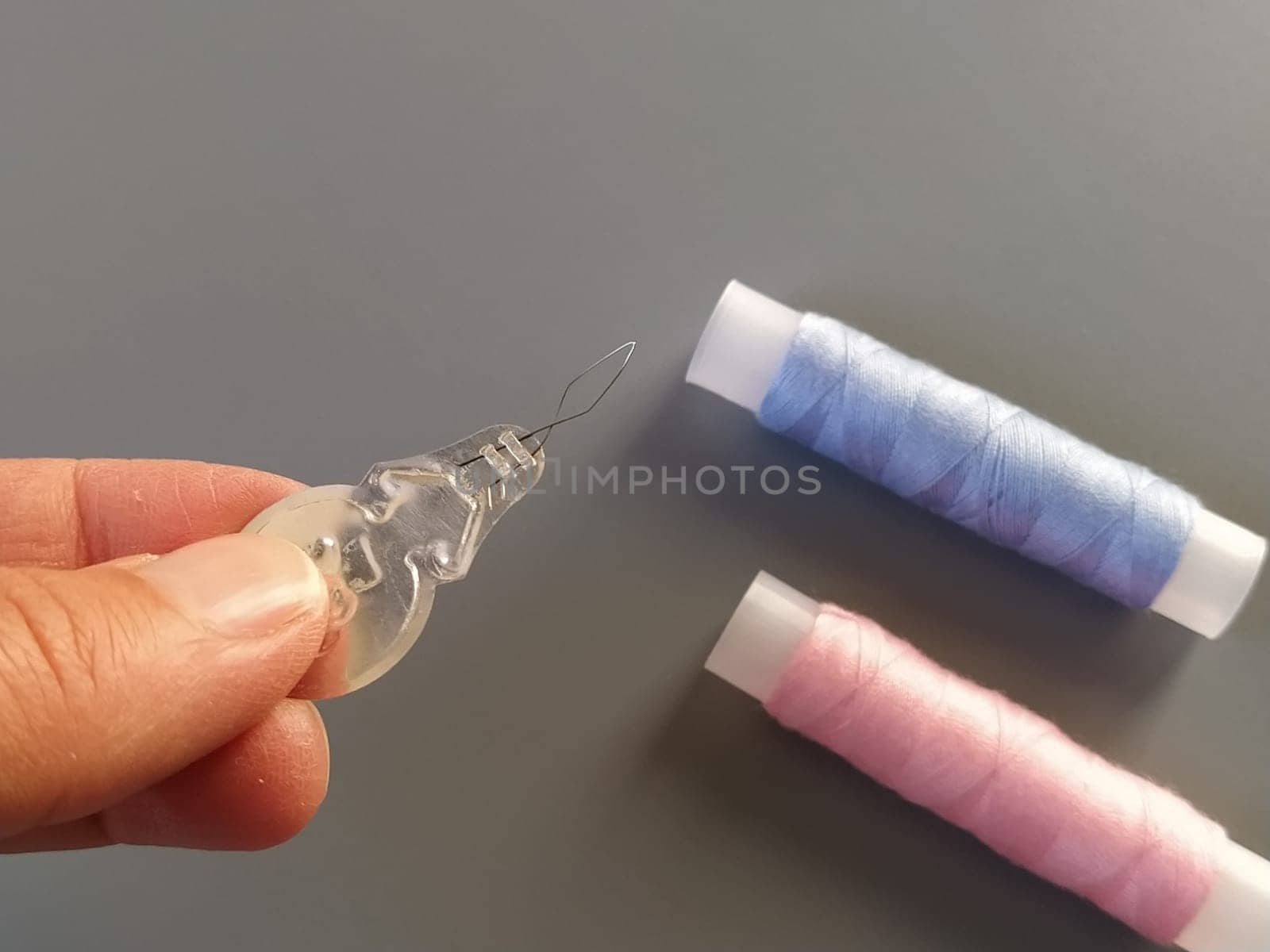 device for drawing the thread into the eye of the needle. Bobbins with blue and pink threads in the background. A woman holds a needle threader in her hand and shows its thin wire eye.
