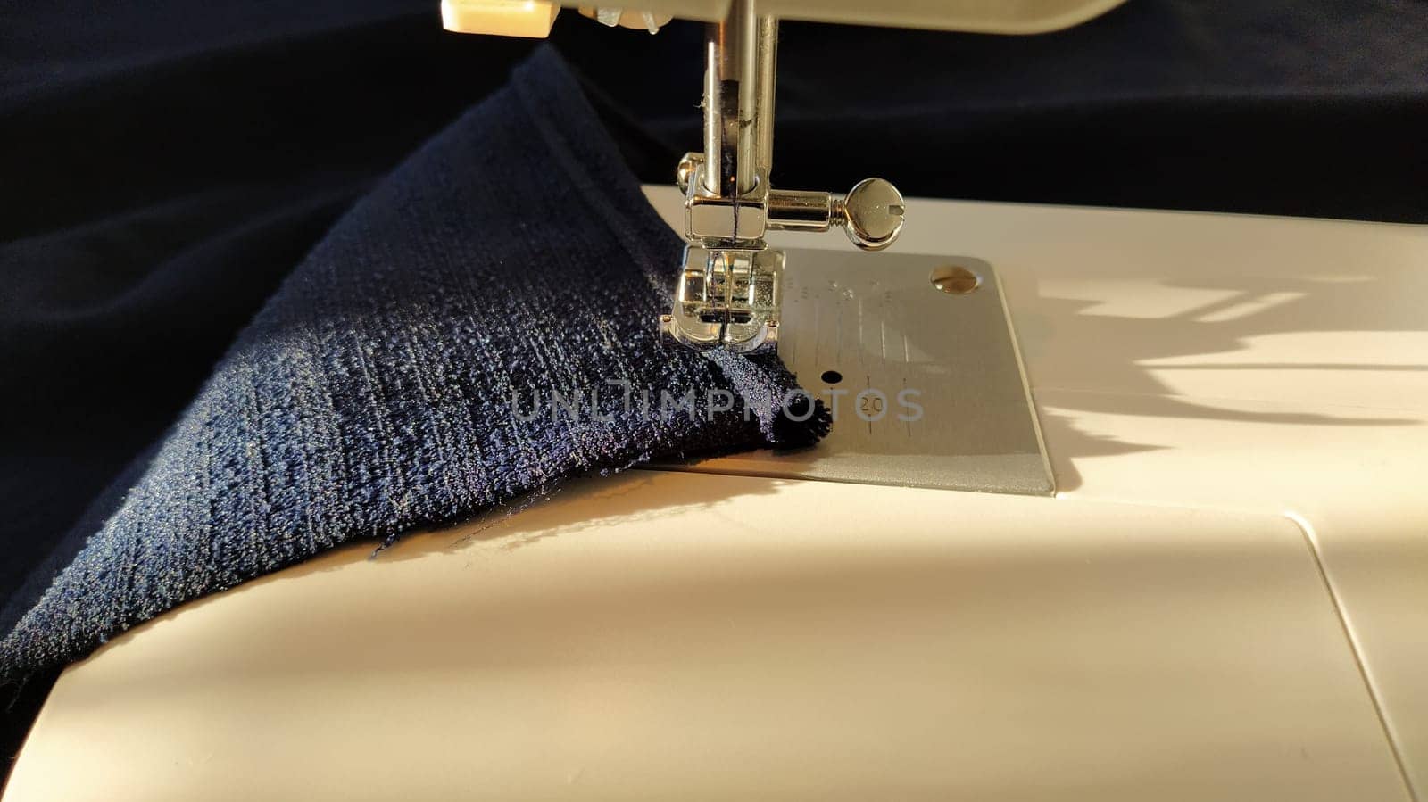 Sewing process on a modern sewing machine, close-up. Sewing machine foot with a needle. Dark blue fabric is trimmed with double wrapping of the fabric edge. Natural sunlight.