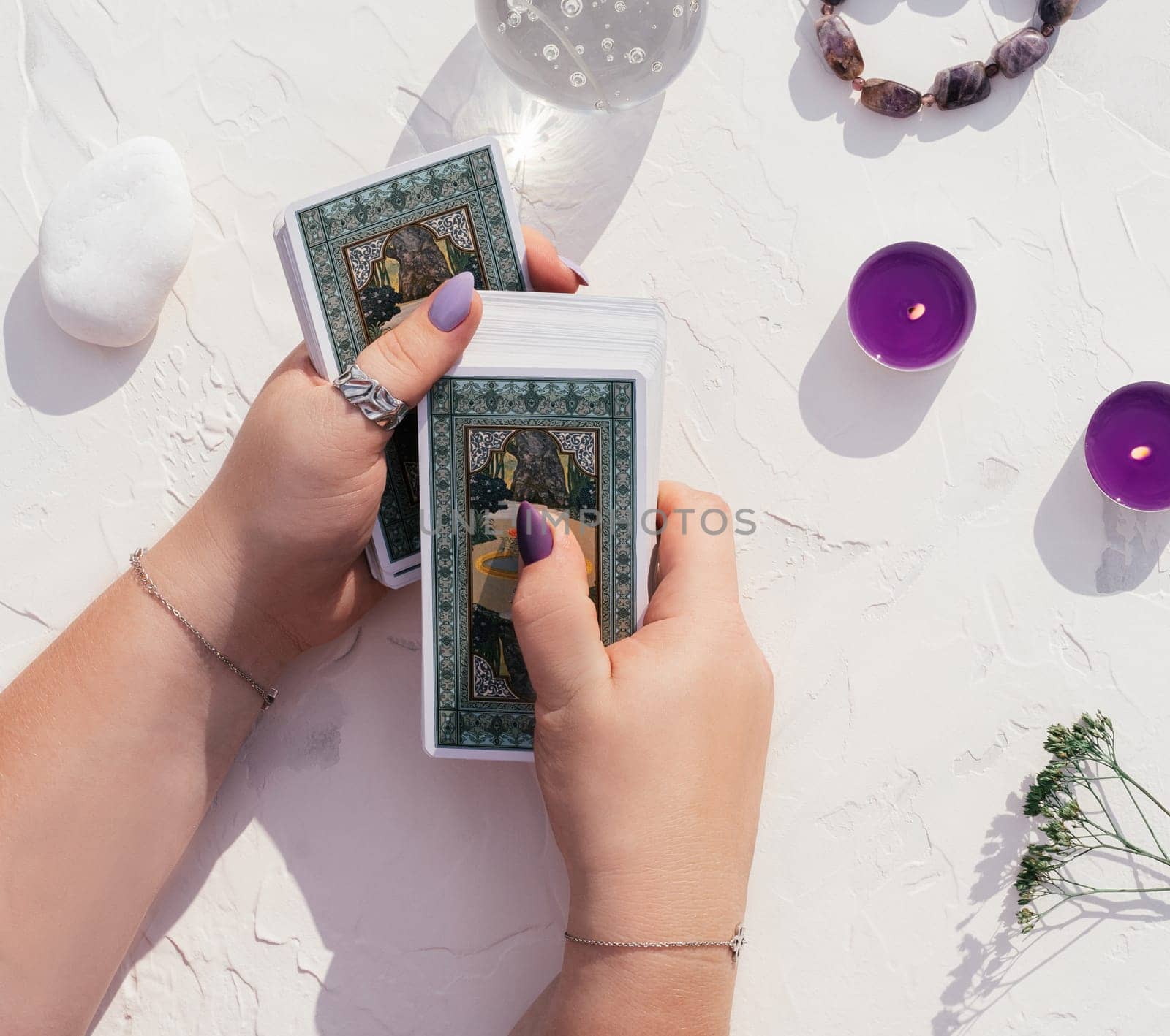 Hands with purple nails and rings hold deck of Tarot cards on white surface with crystal ball, candles and stones. Top view. Minsk, Belarus - 28.07.2021