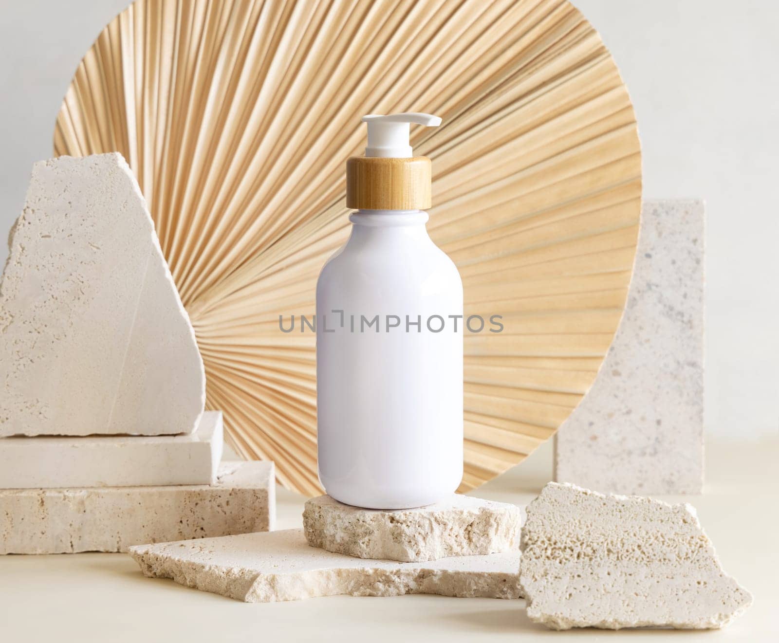 White One pump bottle on biege travertine stones against dried palm leaf, close up, mockup, copy space. Skincare beauty product. Eco friendly cosmetics