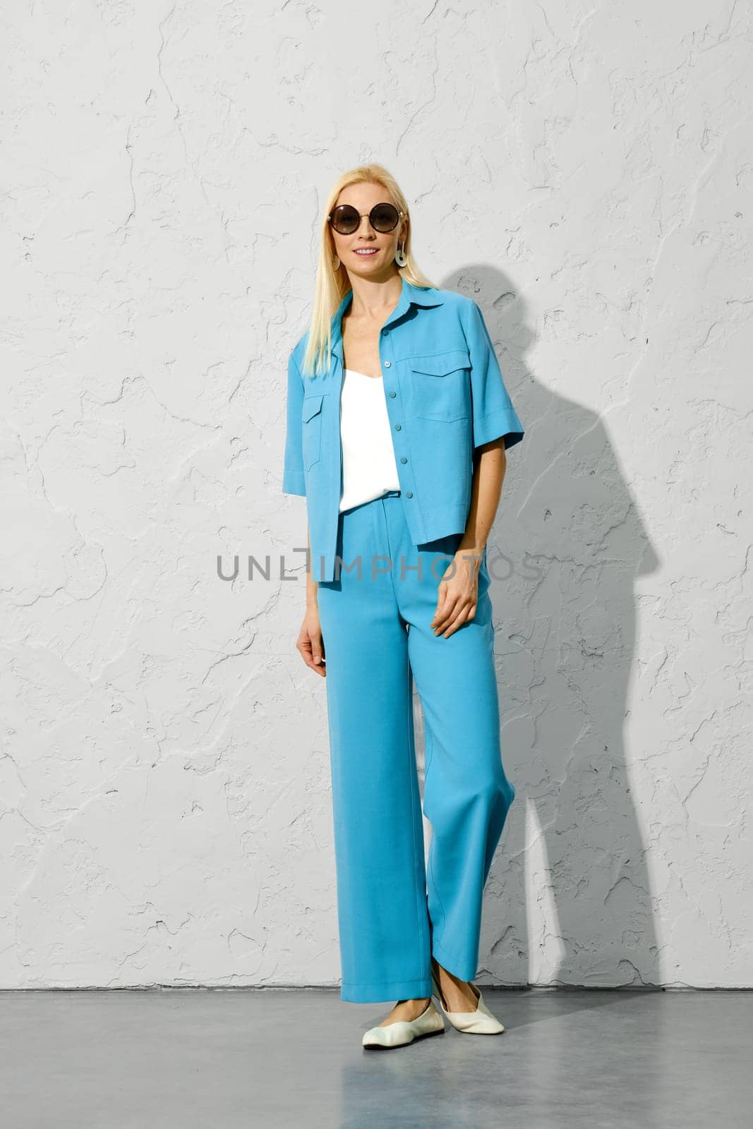 Smiling woman in sunglasses and blue fashionable spring outfit standing near a white wall