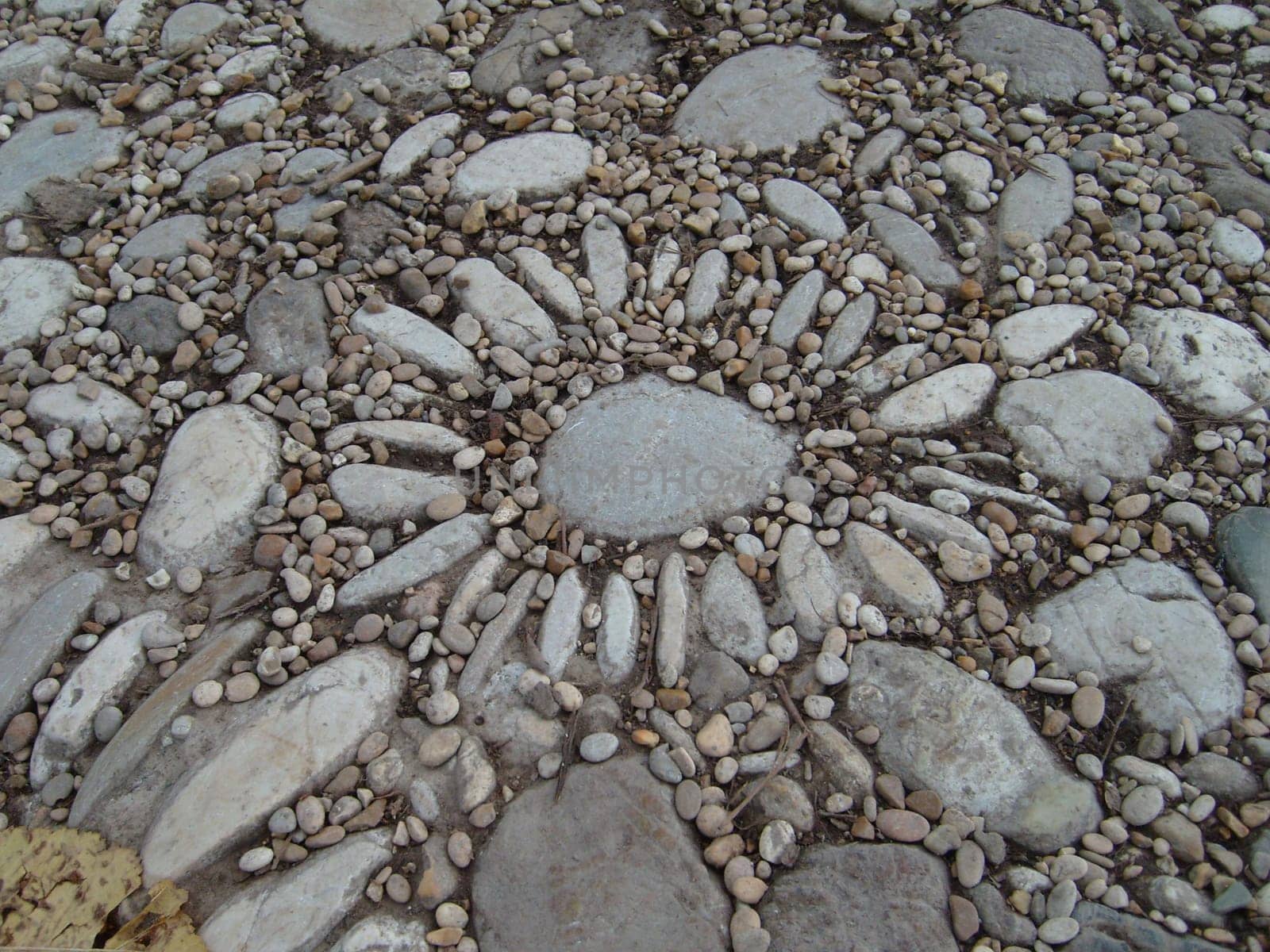 Lined with small and long stones. The pattern on the ground resembles a daisy or the sun. Petals are like rays. Gray small stones neatly laid out in the shape or image of a flower.