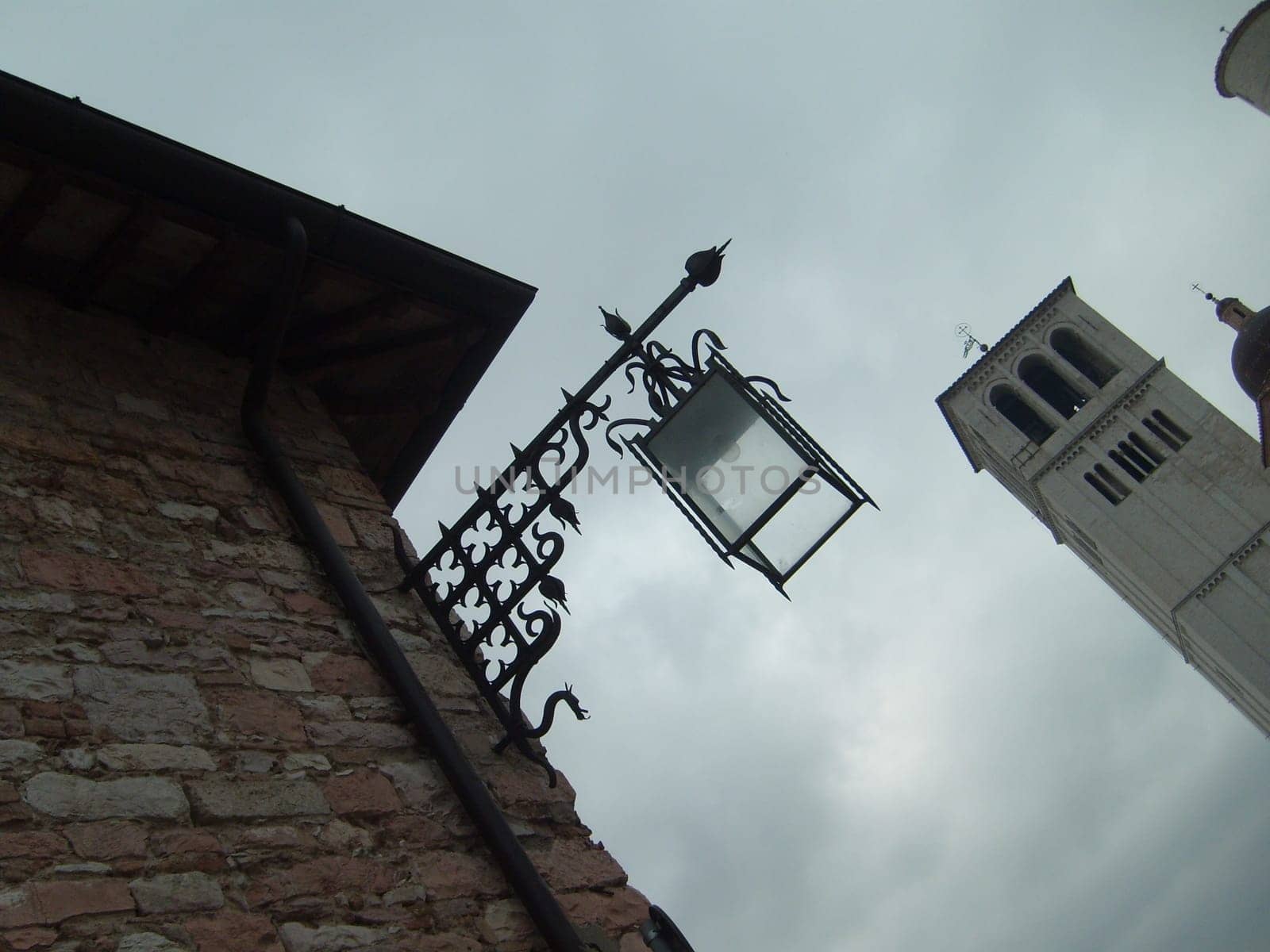 Assisi, Italy, December 1, 2007 Photo taken from an angle. Bottom up view of an old Italian tower and a beautiful metal street lamp. Brick wall of a building with an arrow-shaped lantern