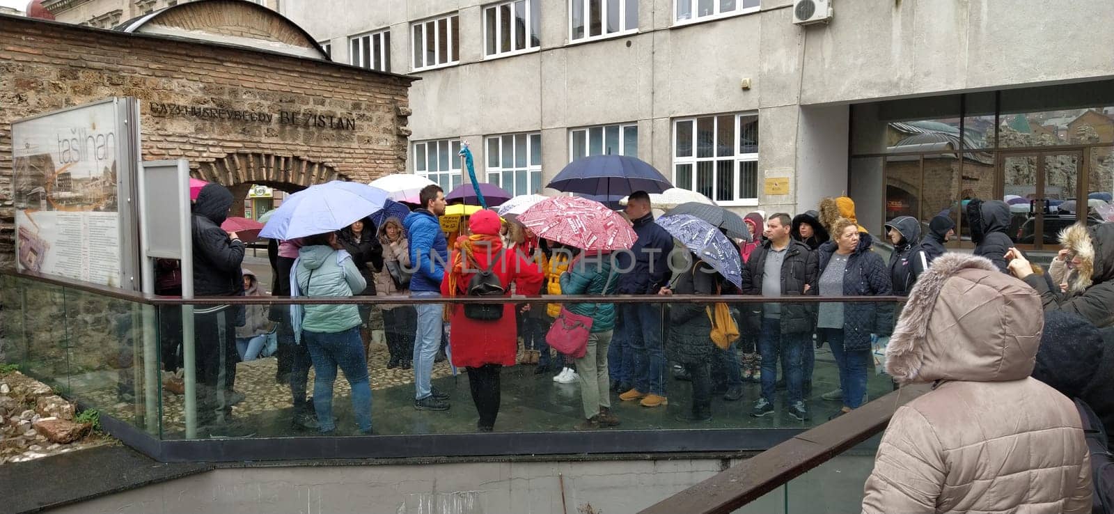 Sarajevo, Bosnia and Herzegovina, March 8, 2020. A group of tourists listening to the guide. It is raining, people are standing under various umbrellas. Ancient buildings made of stone. Bezistan.