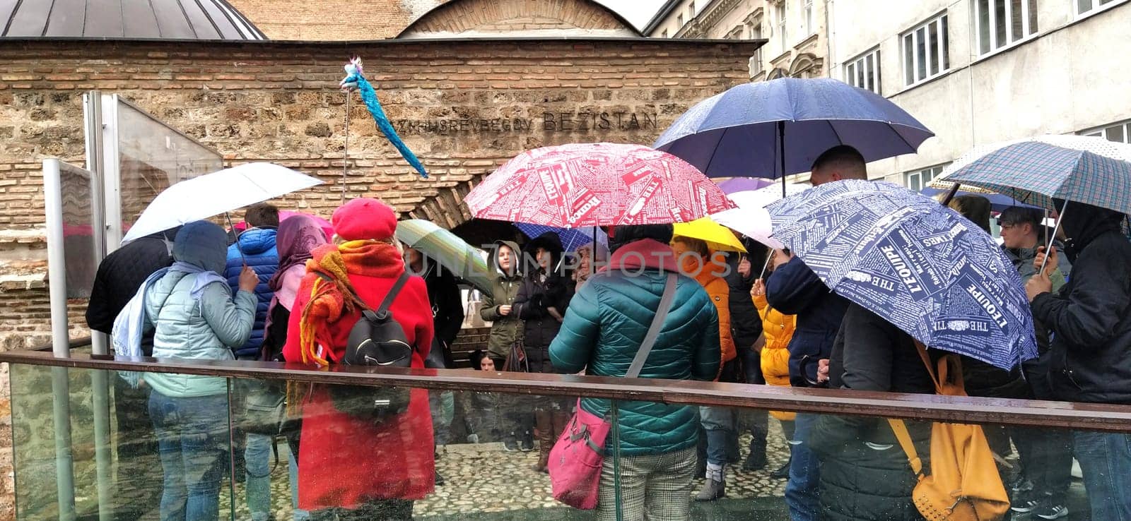 Sarajevo, Bosnia and Herzegovina, March 8, 2020. A group of tourists listening to the guide. It is raining, people are standing under various umbrellas. Ancient buildings made of stone. Bezistan.