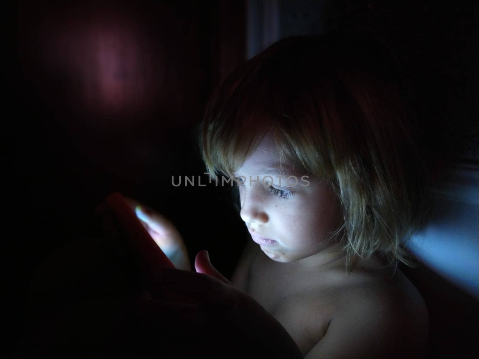 Cute little girl plays with a tablet computer. A child of 7 years old with a serious face and blond hair looks into a gadget. Copy space. Girl in the room. Dark and light wall. At home in the night.