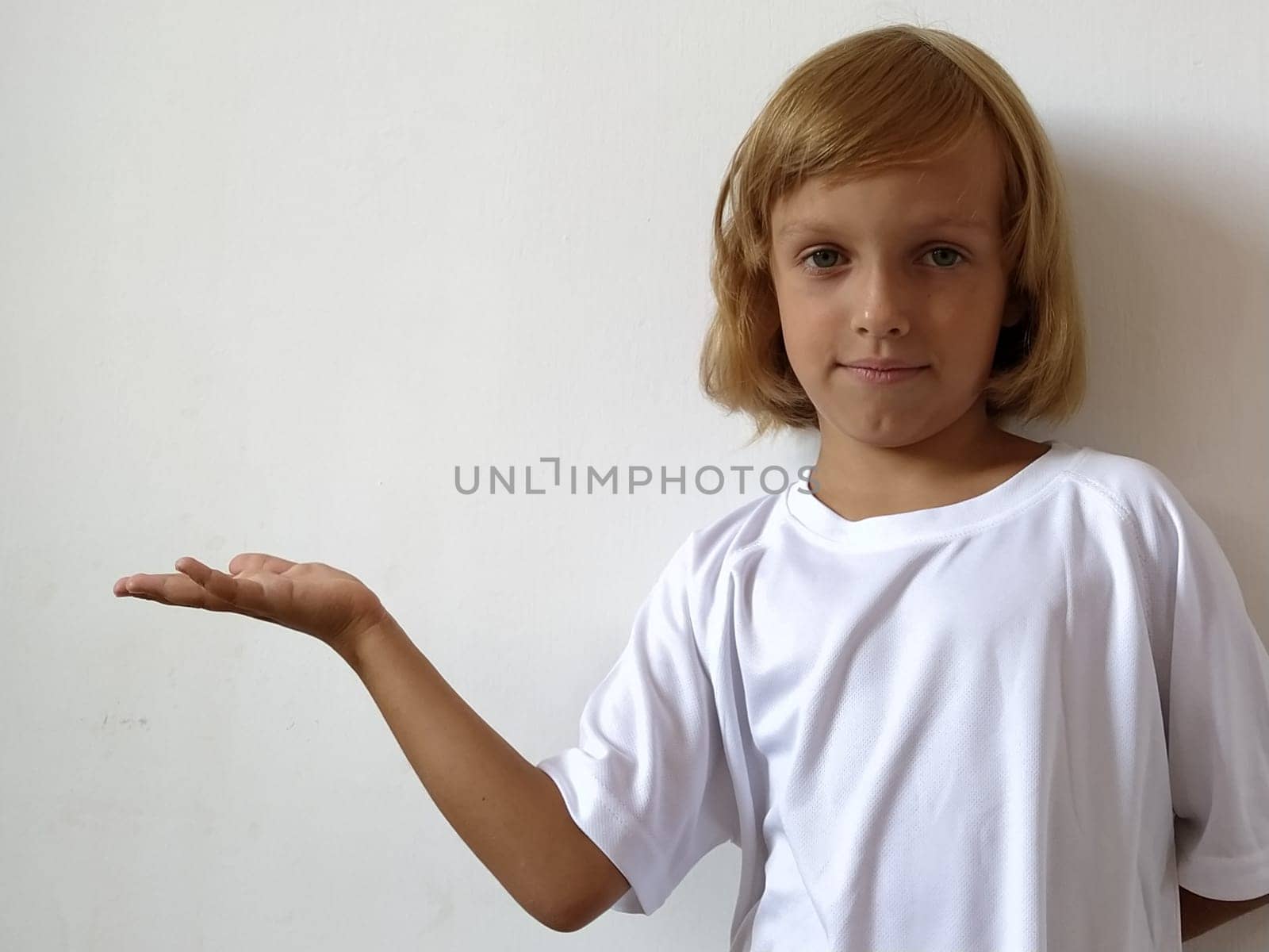 Girl with her hand points to something. Copy space. A little cute girl 6-7 years old with blond hair expresses emotional joy and a smile on a white background. A child with tanned skin. White T-shirt.