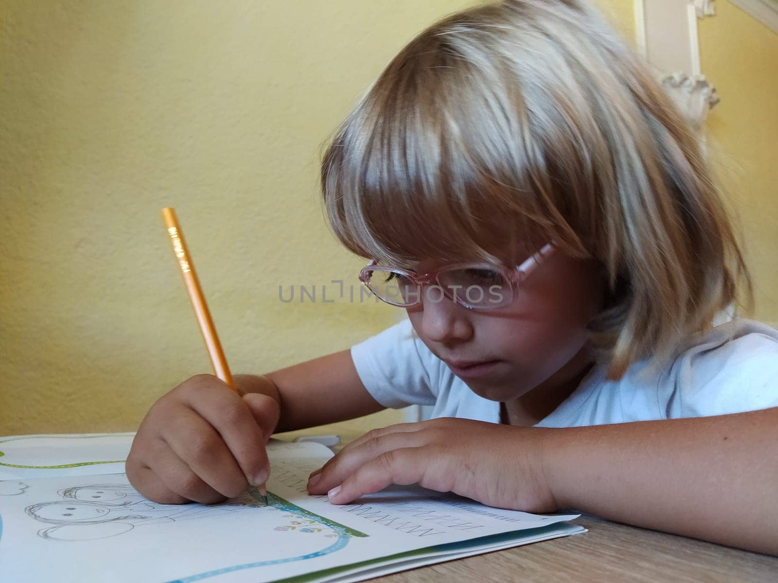 A 6-7 year old girl is reading a book or textbook and smiling. The child has blond hair, dark skin and glasses on his face. The child sits at the desk. yellow wall in the background.
