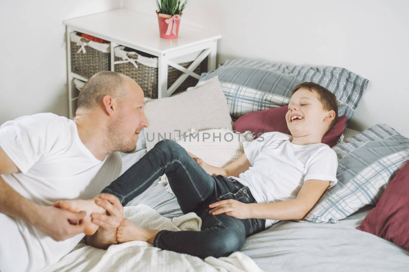 Father plays with his son 7-10 on bed. Dad tickles kids feet. Family having fun