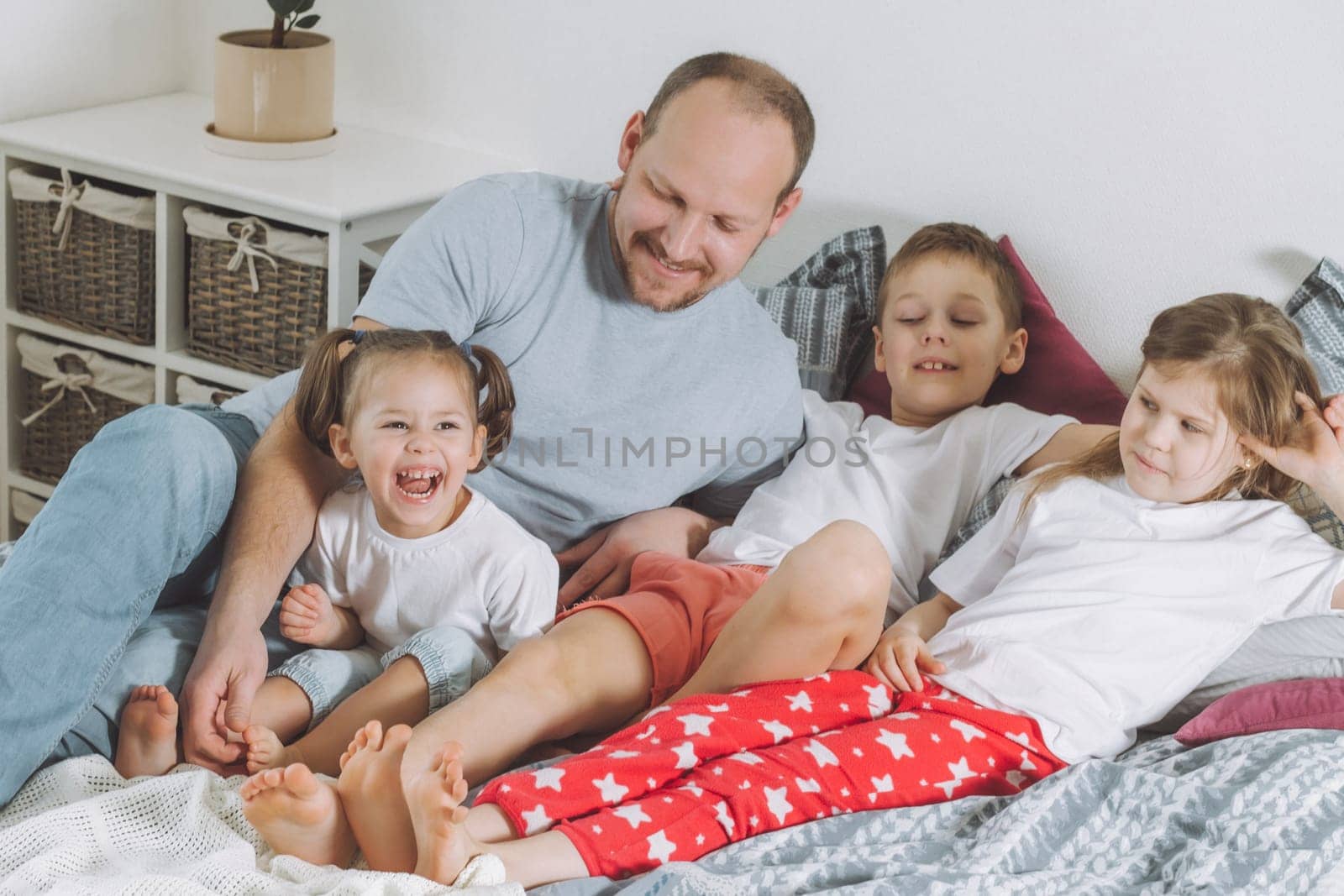 Father plays with three children on bed. Dad tickles kids feet. Family of daddy, two girls and boy