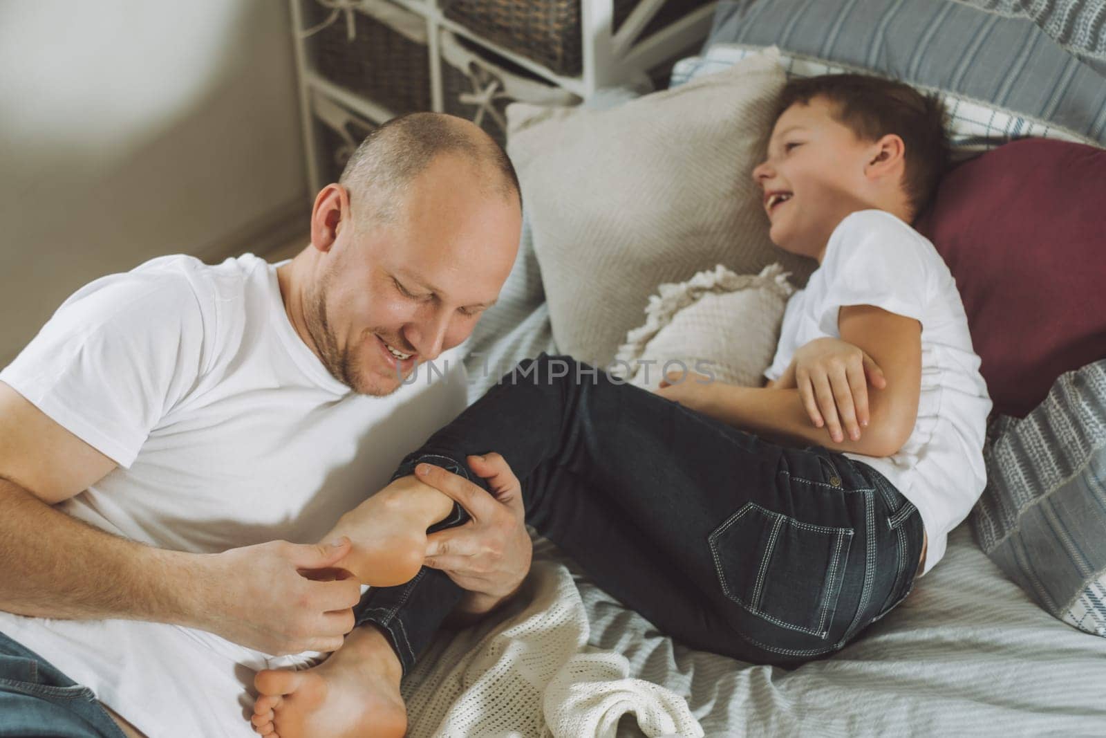 Father plays with his son 7-10 on bed. Dad tickles kids feet. Family having fun