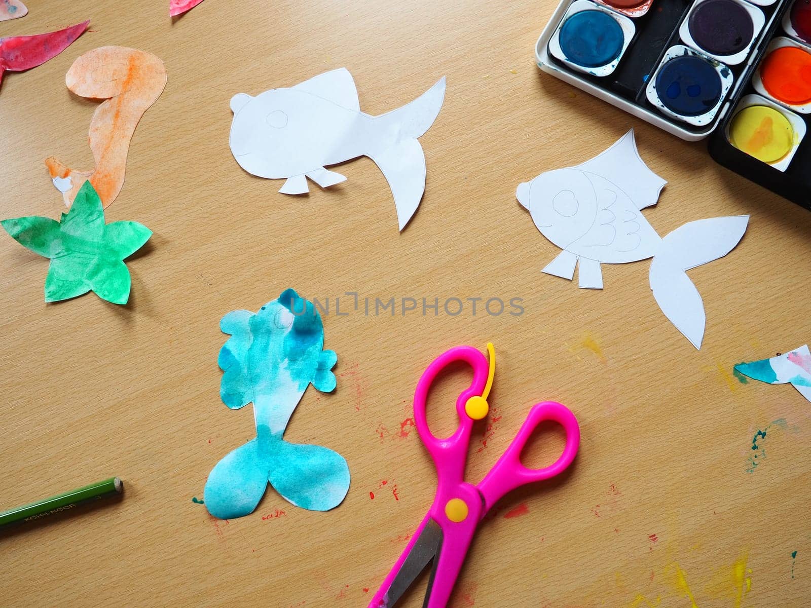 Desktop with drawing tools. A set of watercolor paints, pink scissors, pencil. Paper-cut, painted fish figurines. View from above. Lesson or creative task.