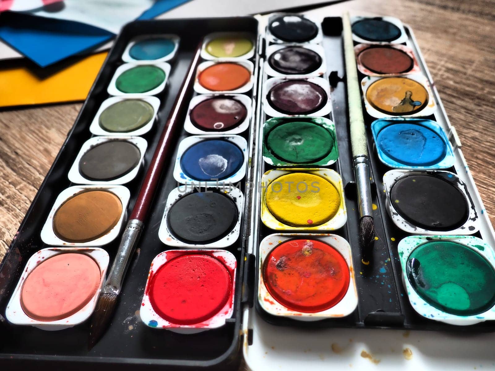 A set of watercolors of different colors for drawing. Multicolored round containers with bright paint. Tools for the lesson of drawing and labor. Colored paper. Red, blue, yellow, green colors