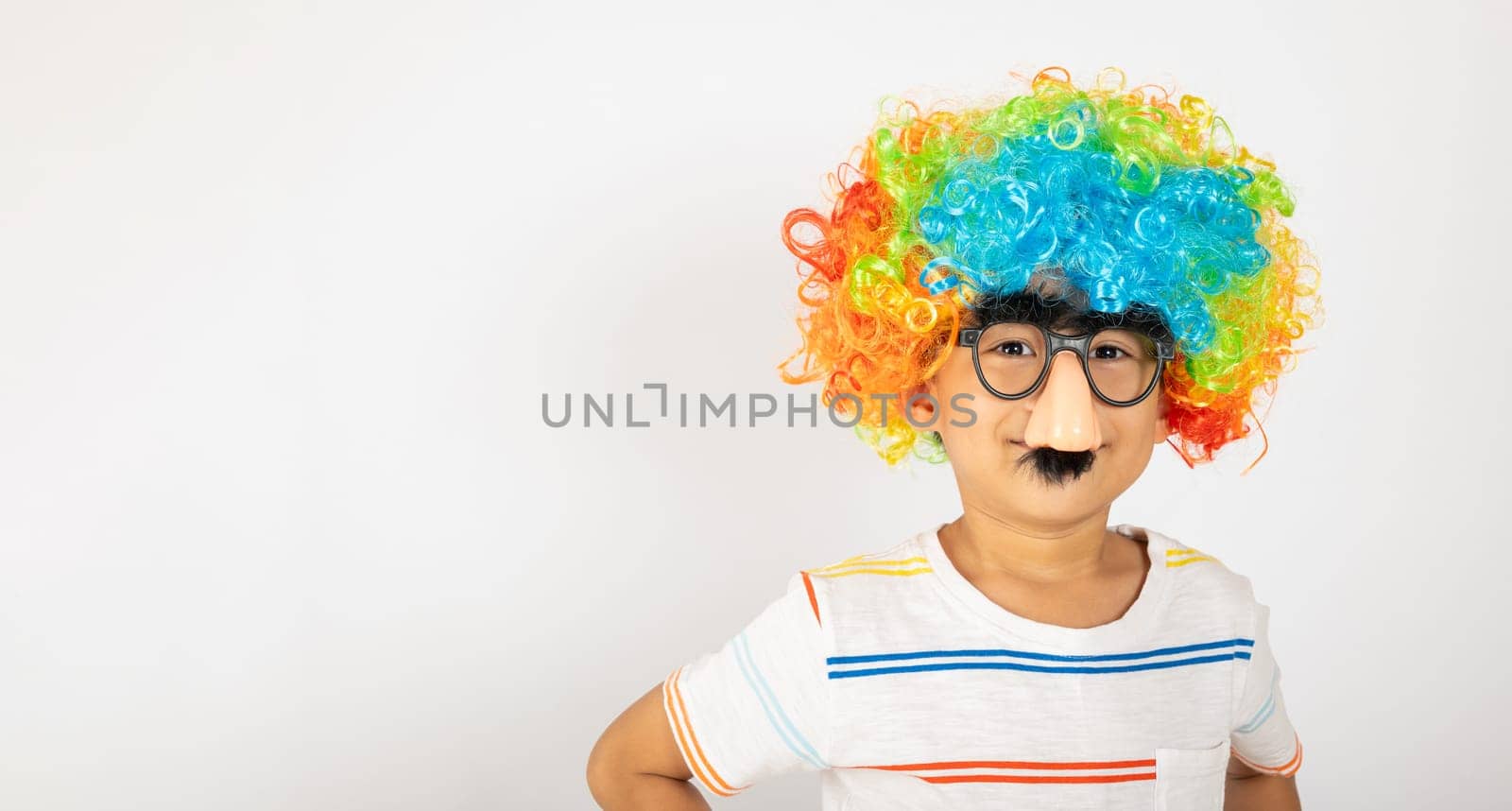 April Fool's Day. Portrait of Funny kid boy clown wears a curly wig colorful a big nos and glasses and has a mustache isolated on white background with copy space, Happy child festive decor