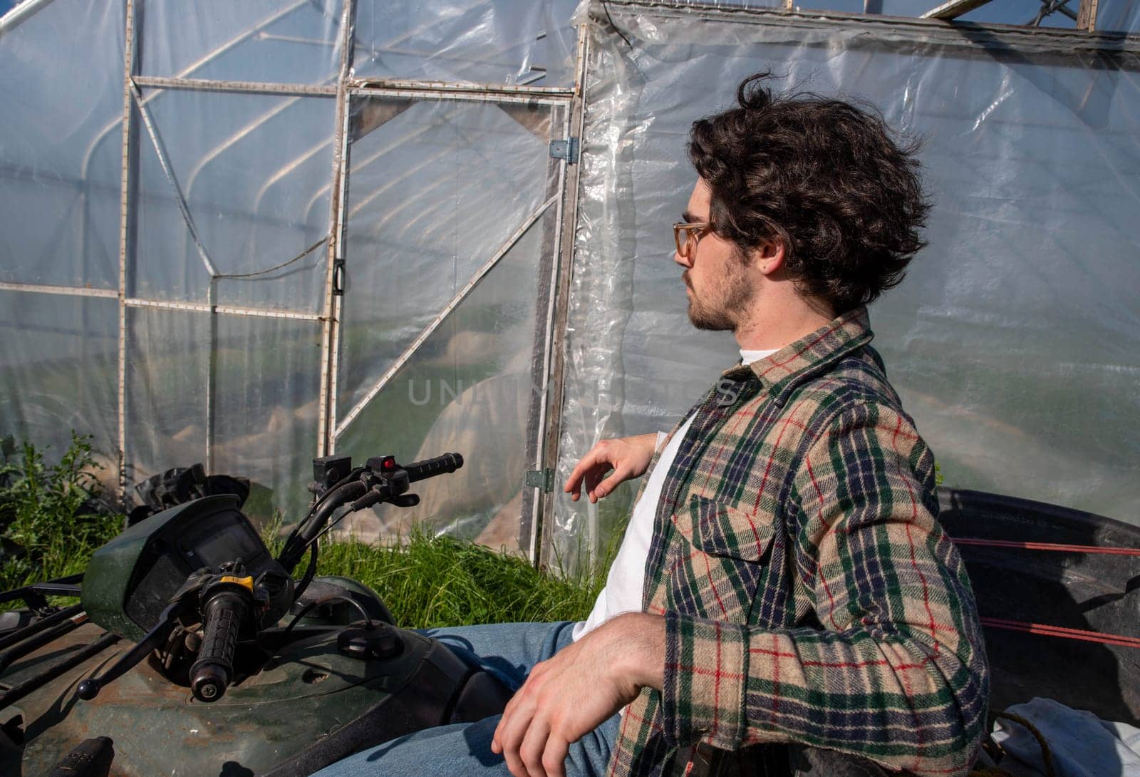 Young caucasion male farmer on an off road farm vehicle contemplating beside an organic vegetable garden greenhouse in natural golden hour sunlight with copy space.