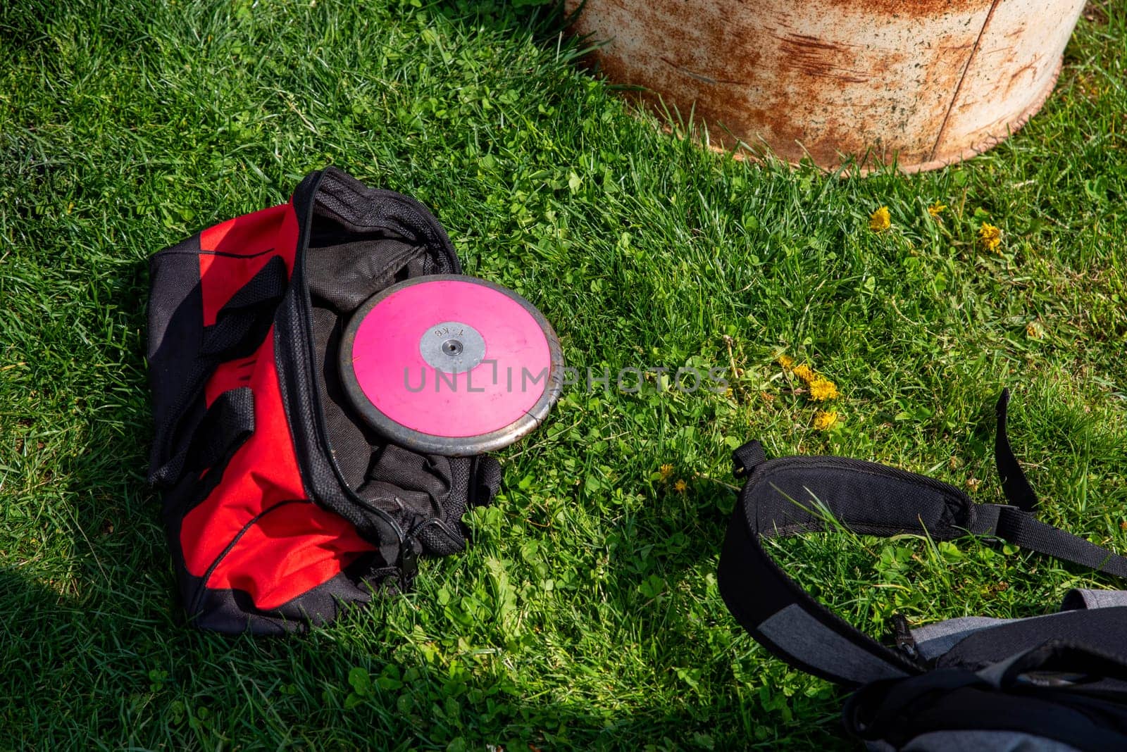 A 1 kg round pink track and field discus in a red athletic equipment bag lays off center in the grass. Shot in natural sunlight with no people and copy space.