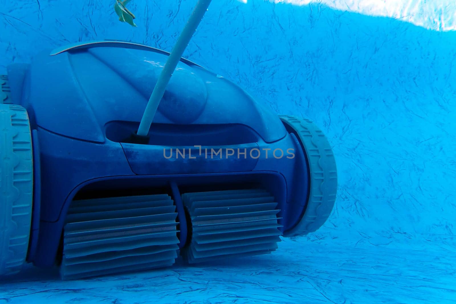 Residential blue vinyl pool liner being cleaned by a motorized wheeled vaccum cleaner with a single leaf in the water. Front wheels and beaters are visible and floating electric cord extends upward. Natural underwater light, no people, with copy space.
