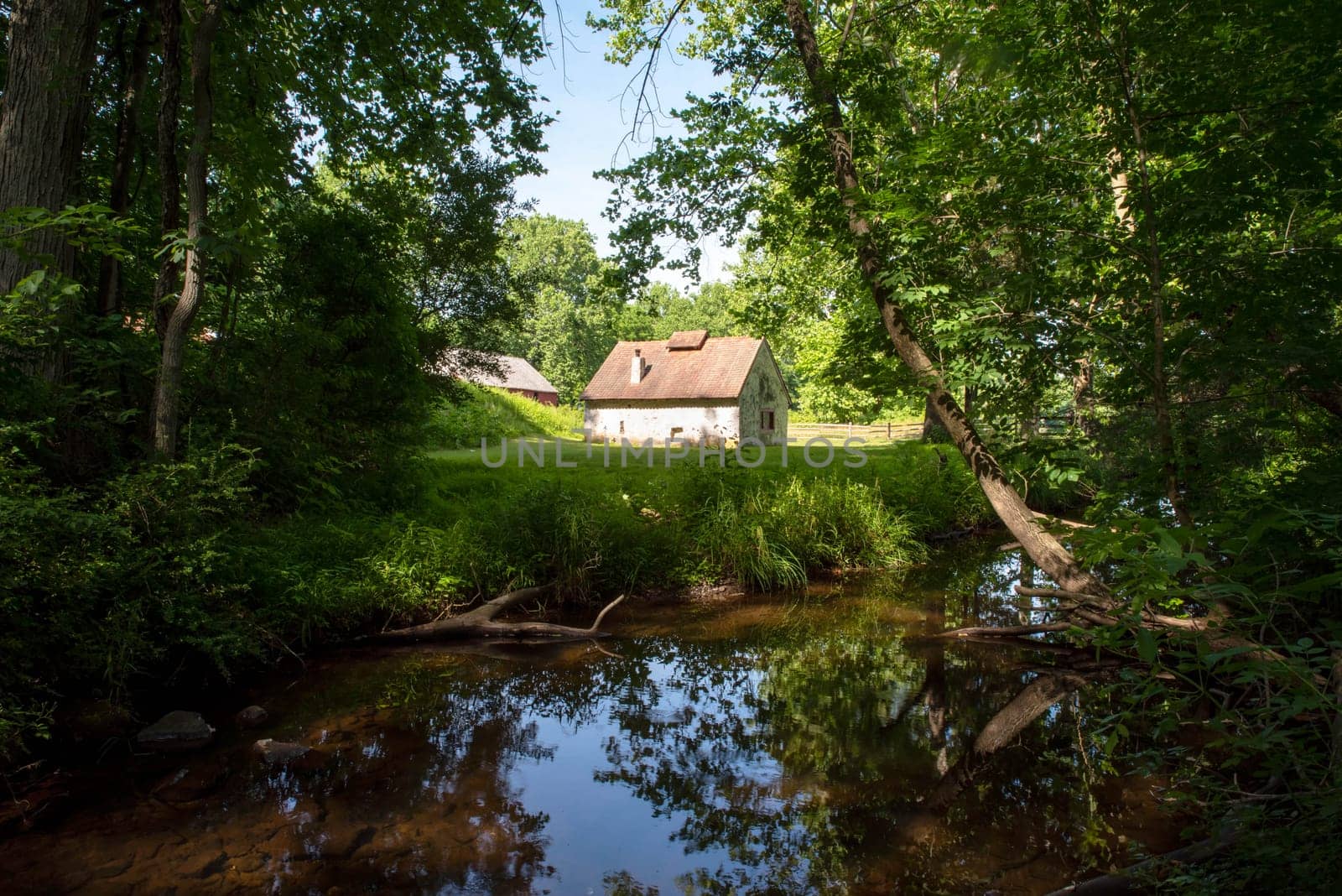 A white stone cottage with a red roof beside a flowing tranquil stream is an enchanted storybook image shot in natural light with copy space and no people. Stream is surrounded by trees.