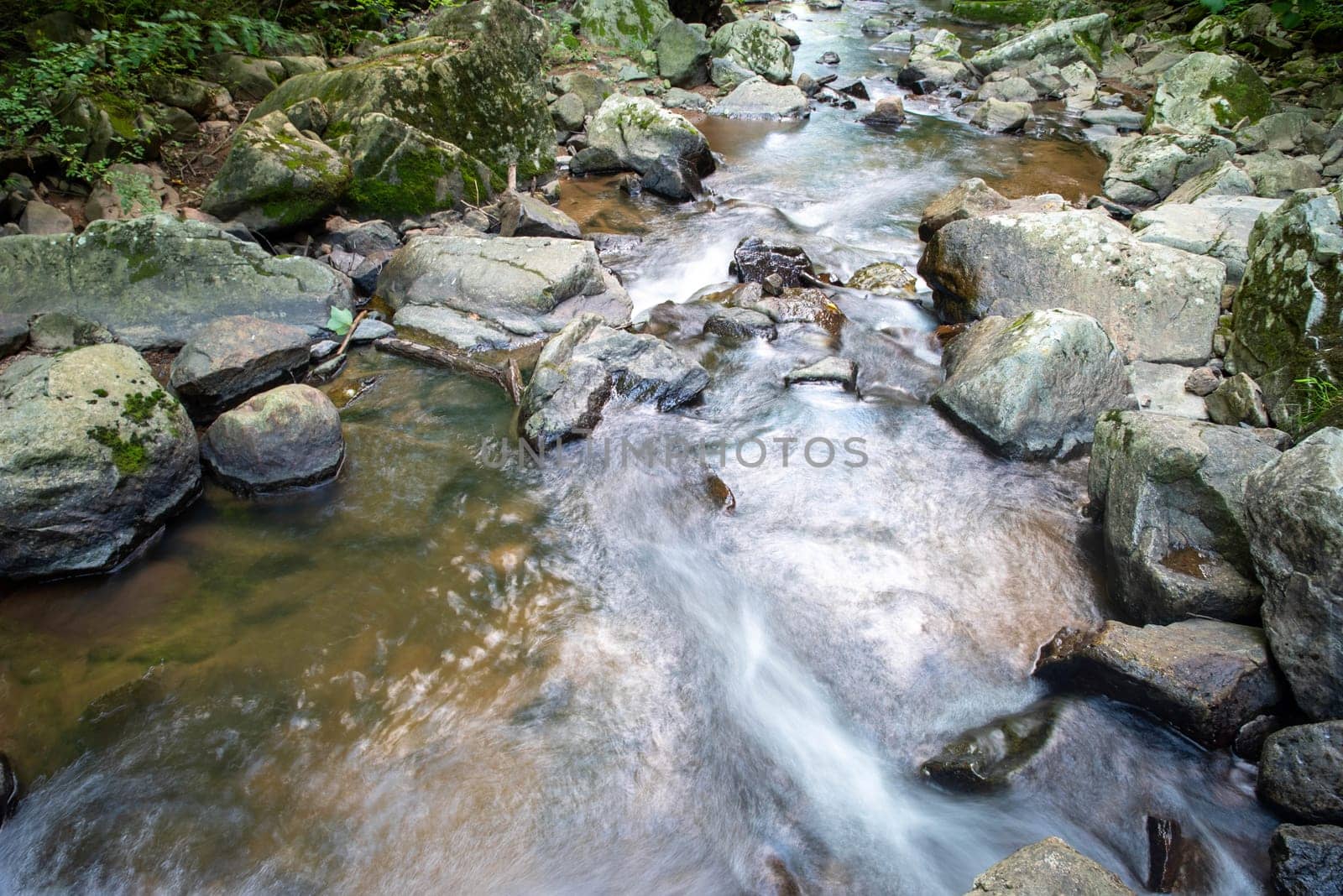 Closeup of long exposure stream flowing around large gray stones and boulders. Water flows away from the camera in a stream nature background. Natural light with no people and copy space.
