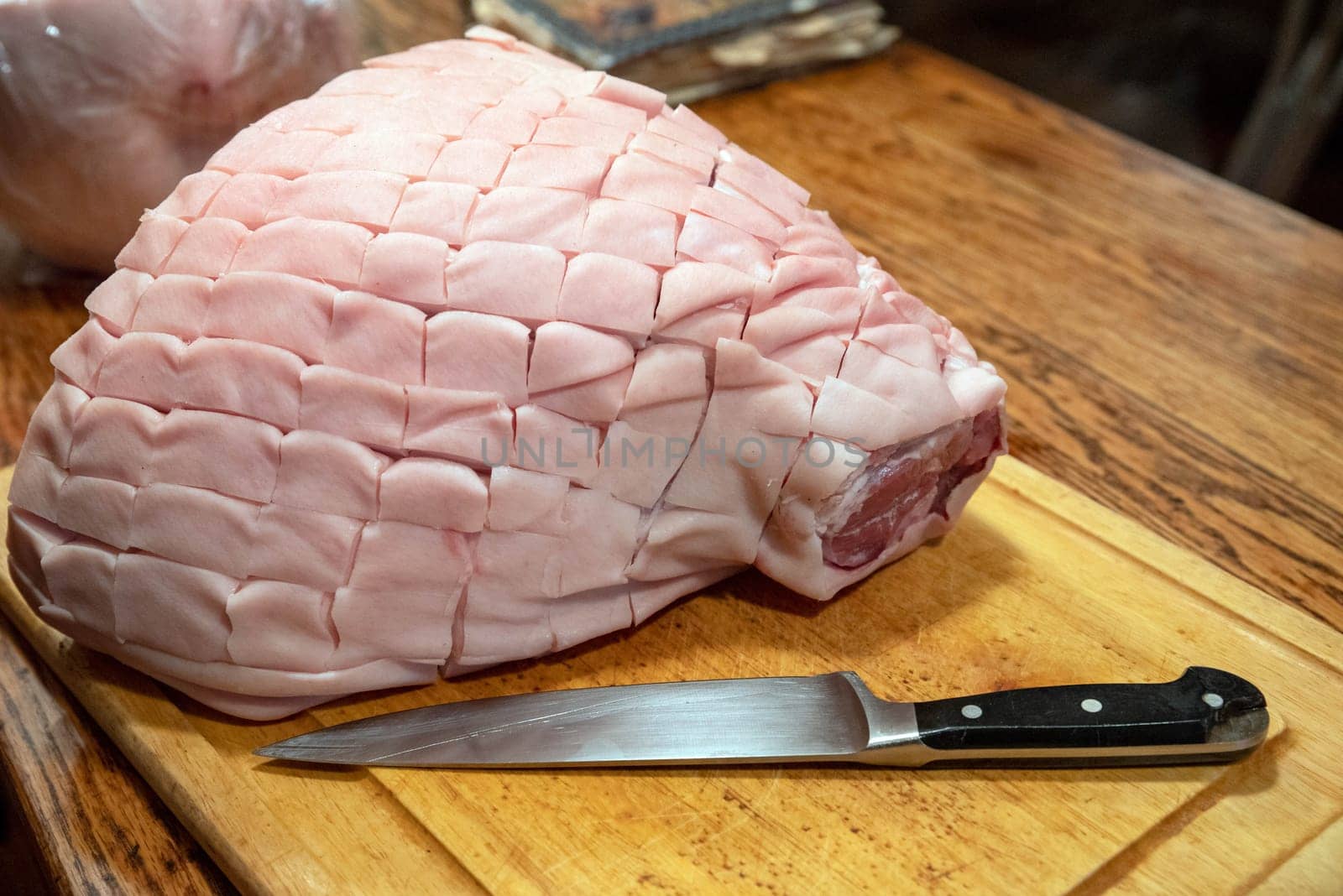 Antique wooden table with wood grain texture and cutting board with black-handled chef's knife and a big skin on bone-in pork shoulder picnic roast. Skin has been cut into diamond cross hatch pattern. No people with copy space