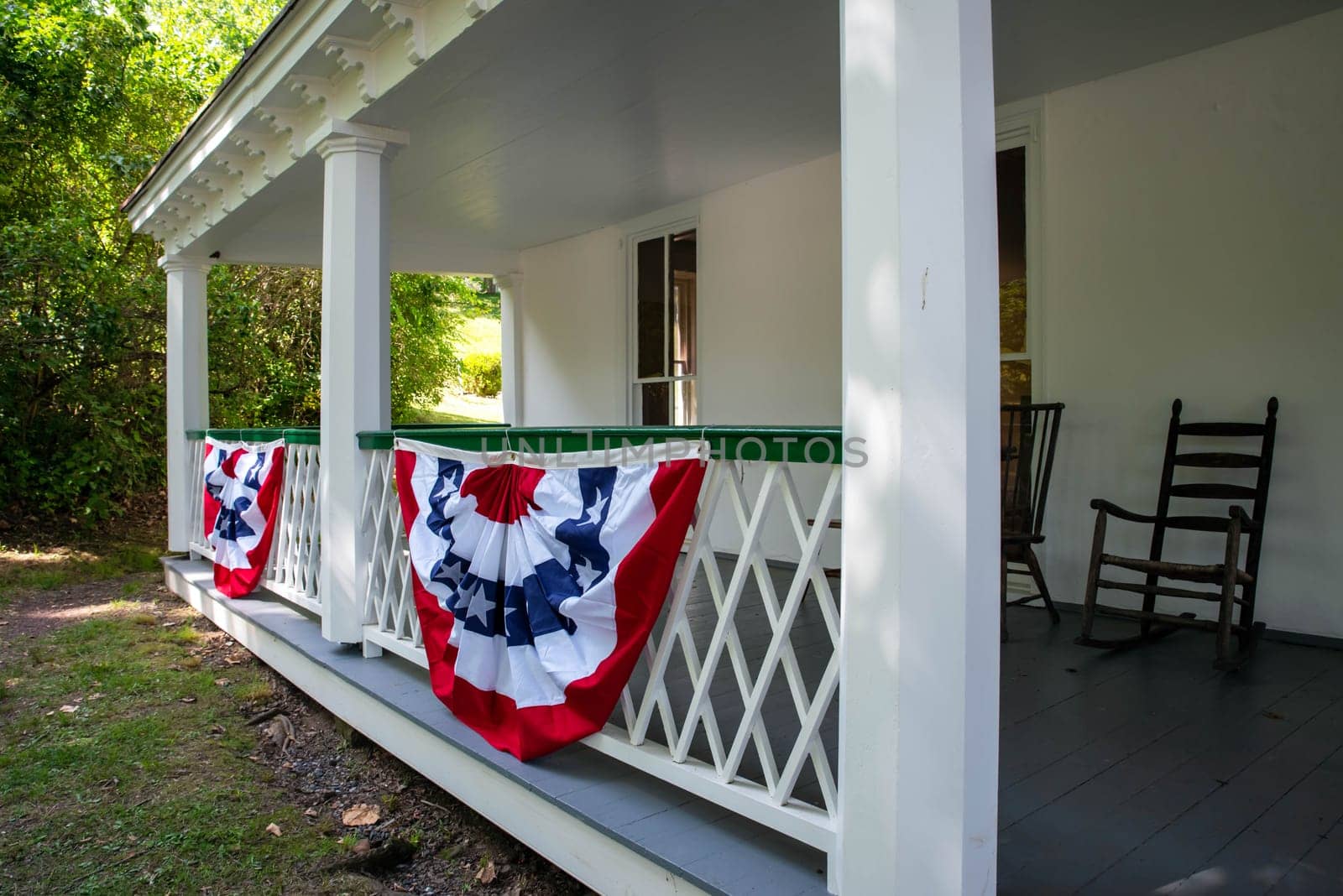Large front porch of an old home draped in old fashioned patriotic American flag holiday decorations with rocking chairs, sunny green grass yard background with no people and copy space.
