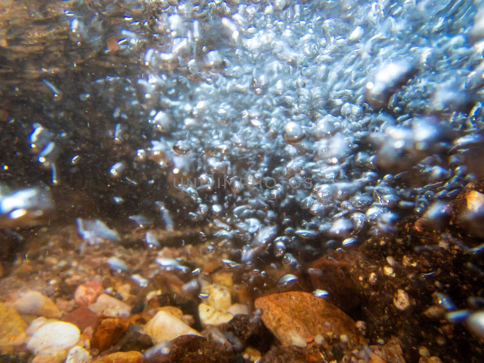 A rush of bubbles swooshes toward the camera in a high energy underwater hydro dynamic image with colorful stones and pebbles on a creek bed. Shot with an underwater camera with flash and natural light, no people, with copy space.