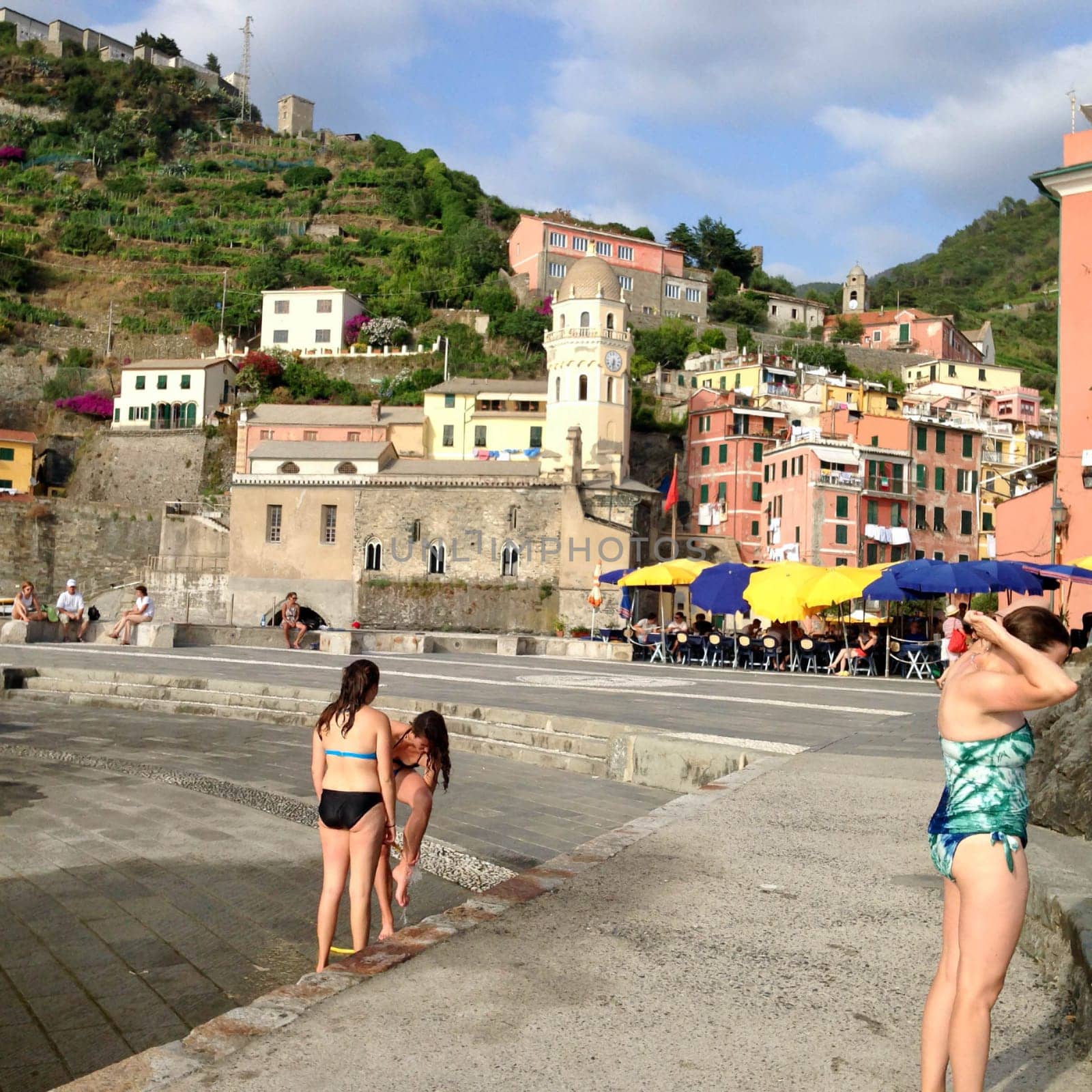 08-05-2014, Vernazza, Italy, Waterfront village scene with church and swimmers Beautiful marina with swimmers and outdoor restaurant with colorful table umbrellas in the foreground of this centuries old village in the Cinque Terre region of Italy. Santa Margherita di Antiochia Church with bell tower and clock as well as ancient houses and green hills on a summer day.