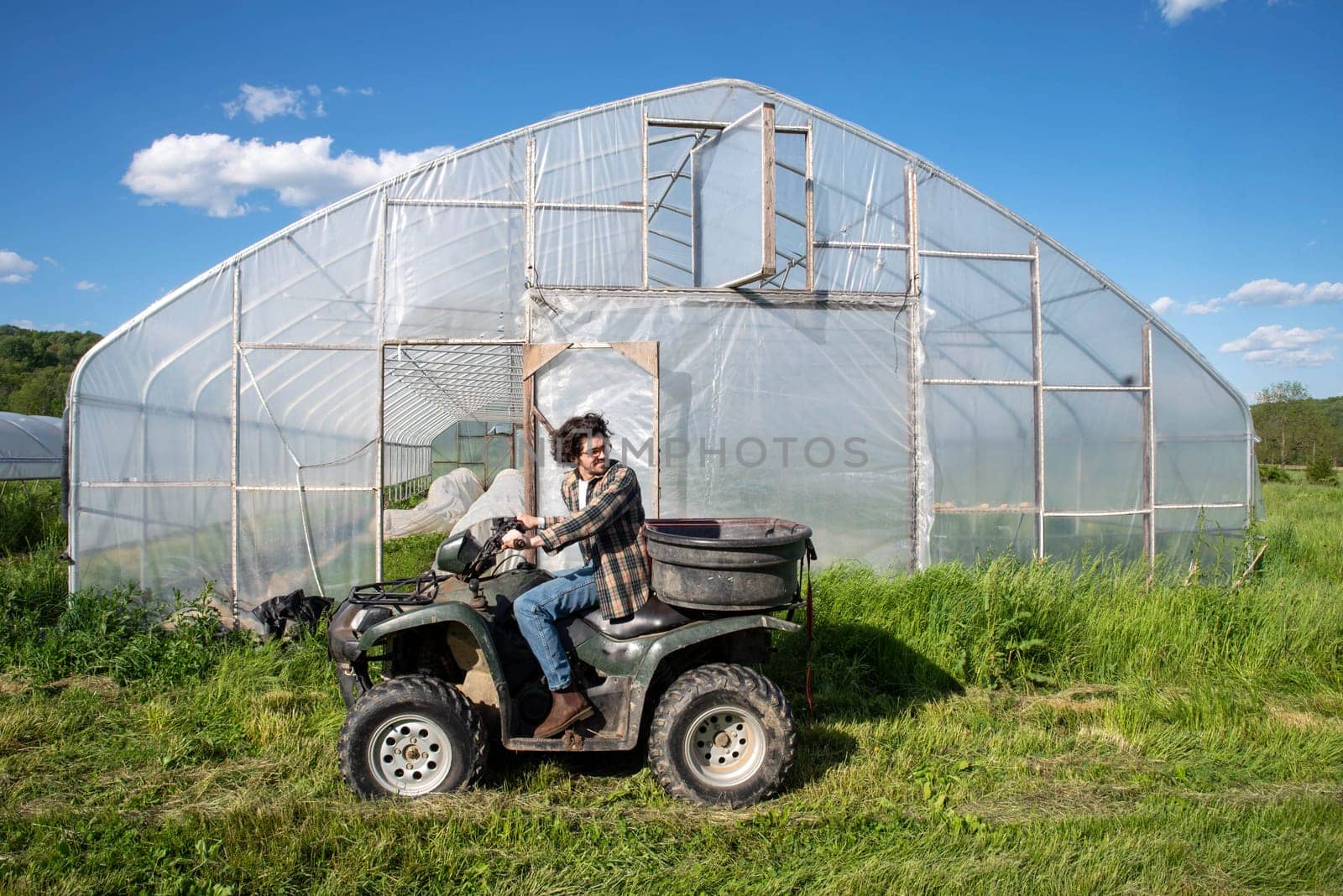 Young caucasian male farmer drives an atv by a vegetable garden greenhouse on an organic farm.Greenhouse door is open with covered rows of vegetables visible inside. Green grass and blue sky, shot in natural light with copy space.
