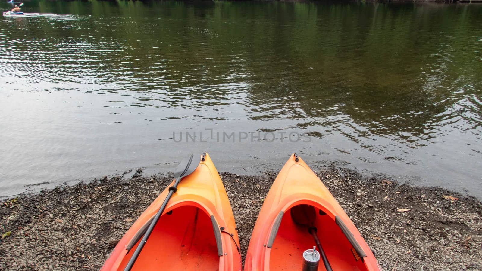 Beautiful outdoor adventure nature background scene with a pair of orange kayaks pointing toward a river and a green woodland scene reflected in the water surface.