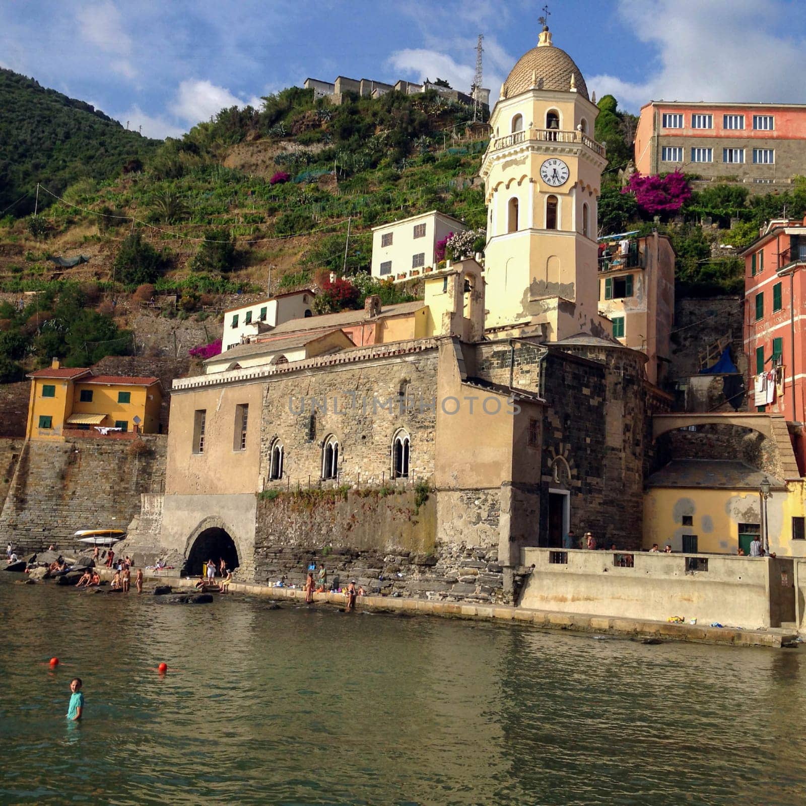 08-05-2014, Vernazza, Italy, Waterfront village scene with church and swimmers Beautiful marina with swimmers and boats in the foreground of this centuries old village in the Cinque Terre region of Italy. Santa Margherita di Antiochia Church with bell tower and clock as well as ancient houses and green hills on a summer day.