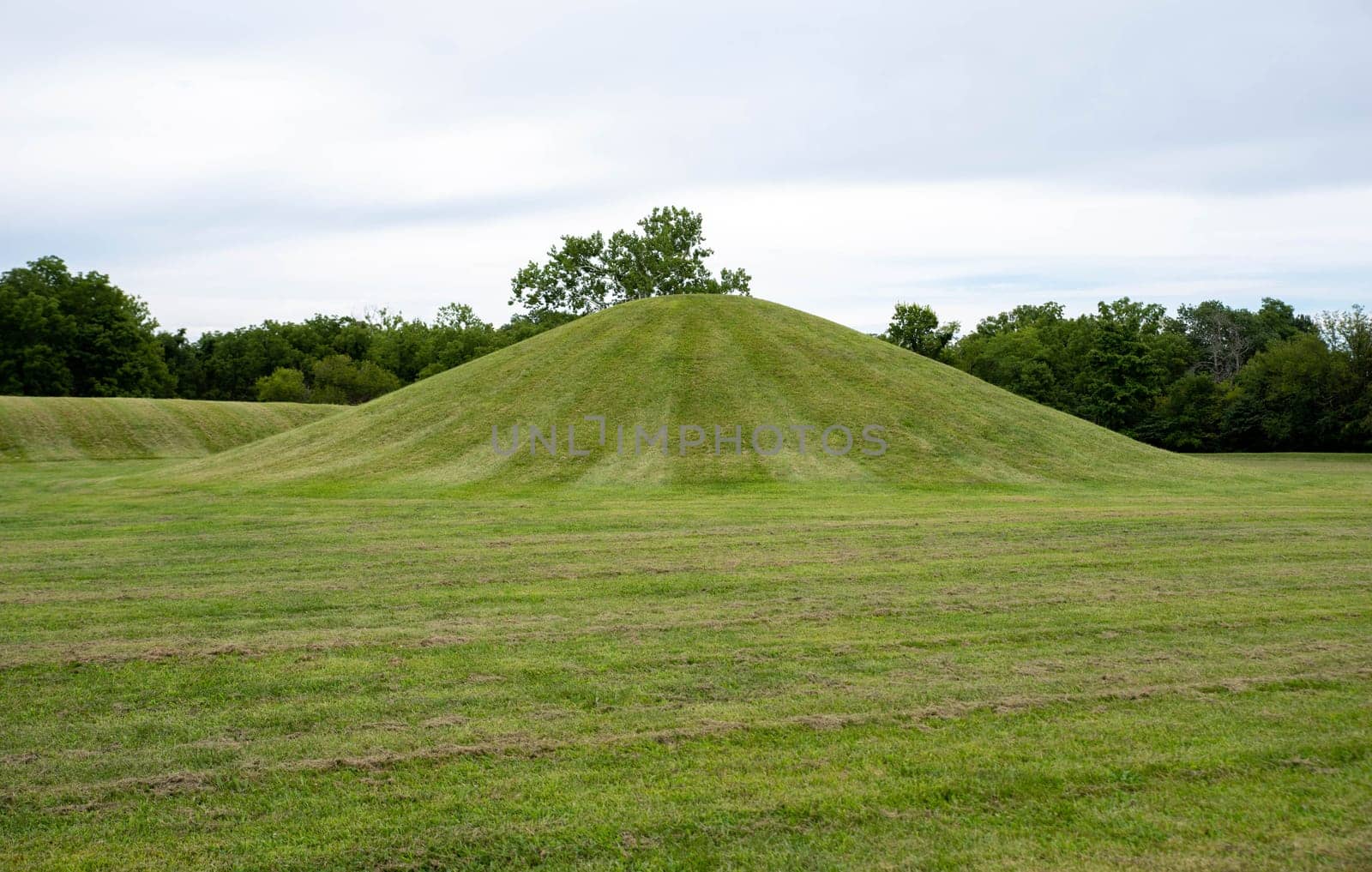Native American Hopewell Culture prehistoric Earthworks burial mounds in Mound City Ohio. Ancient circular mound in foreground and long mound in the background. Grass is neatly trimmed with trees and dramatic sky.